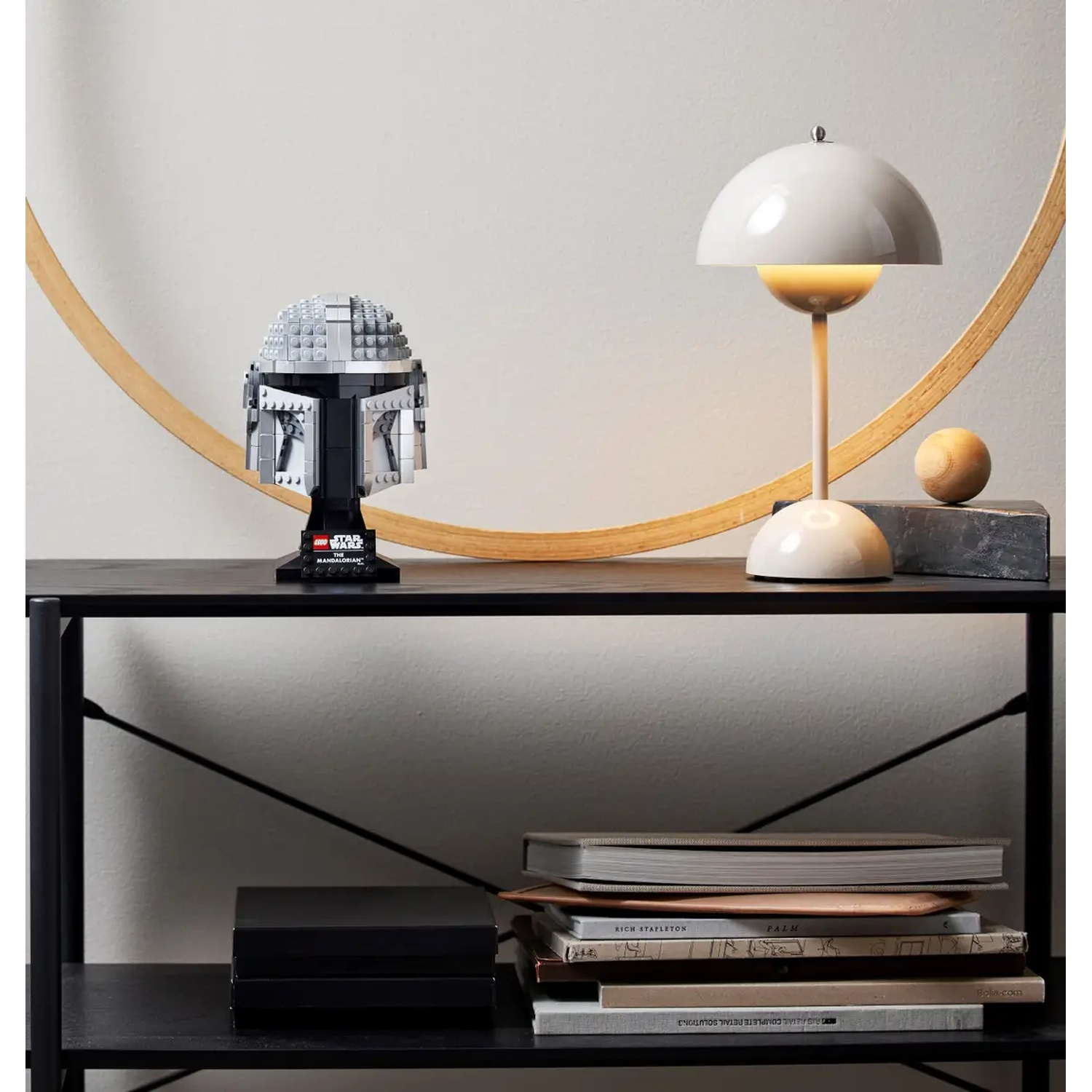Een Mandalorian Lego-helm staat op een plank met een lamp en boeken. Een gouden boog staat erachter. De plank is zwart.