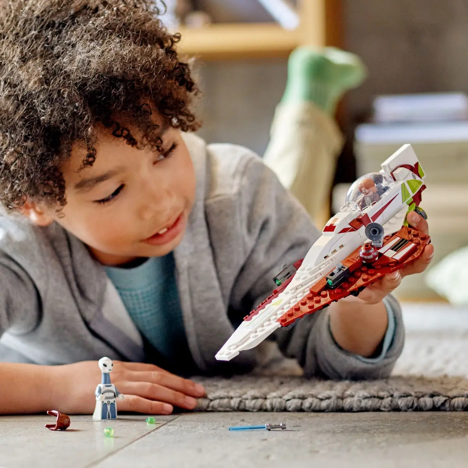 Een kind houdt een LEGO starfighter vast en bekijkt het. LEGO figuren en accessoires liggen verspreid op de vloer. Achtergrond bevat een houten plank.