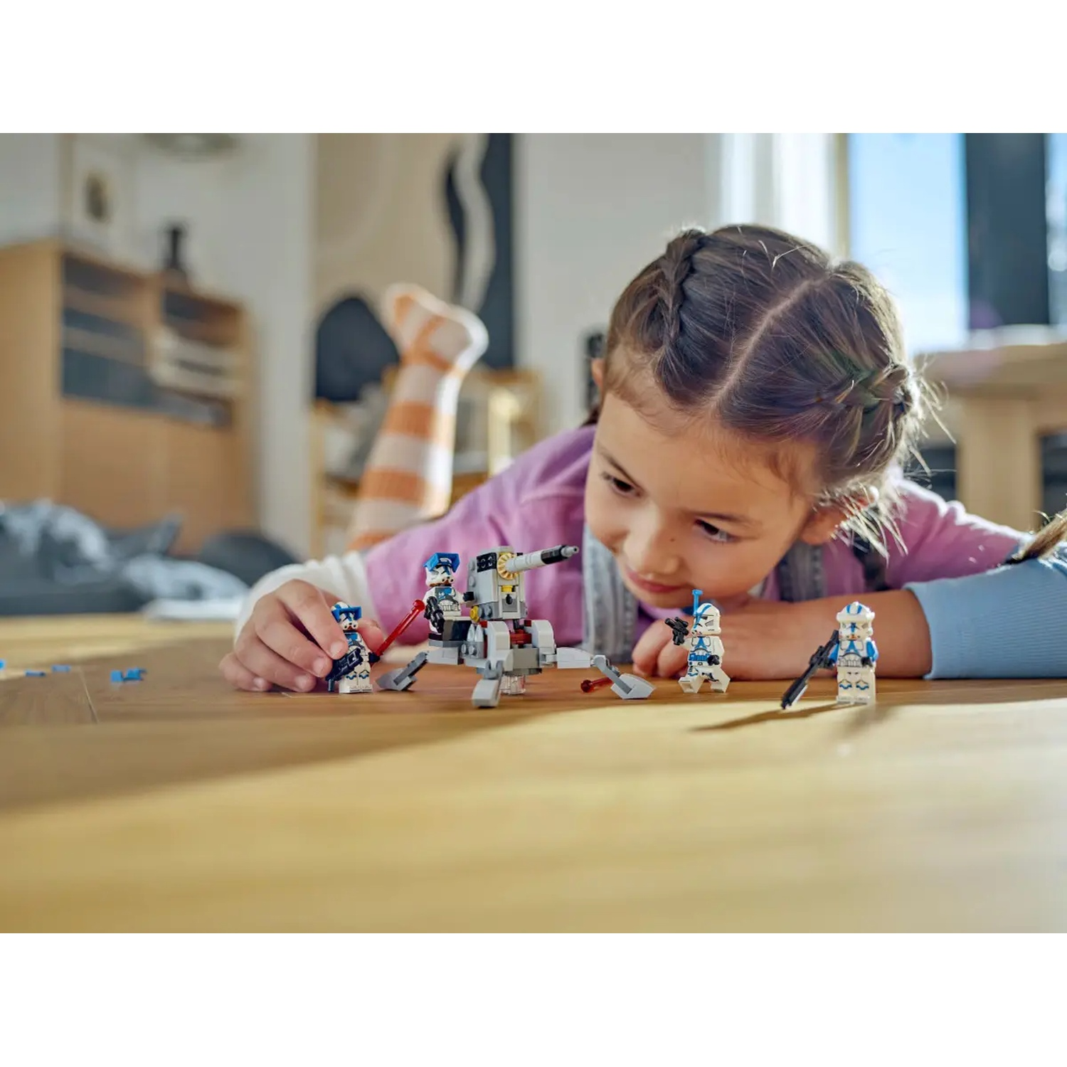 A young girl is playing with Star Wars Lego figurines on a wooden floor. A Lego vehicle and several figures are present.