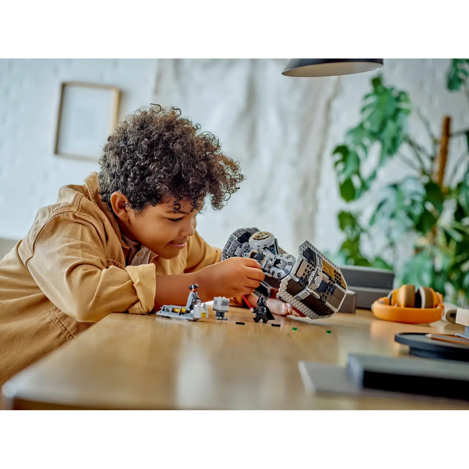 A boy with curly hair plays with a Lego set on a wooden table. The set includes a black vehicle and Lego figures.