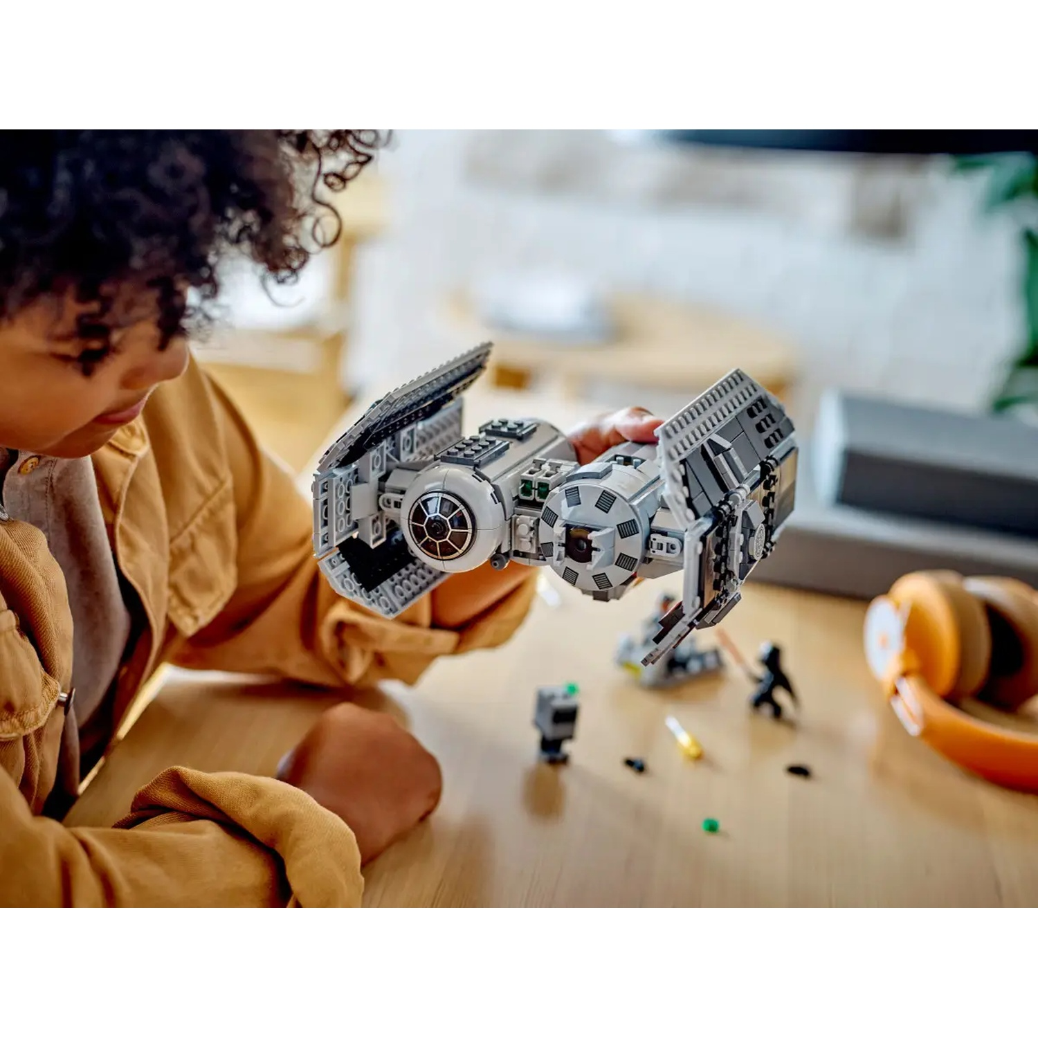 A child holds a LEGO Star Wars TIE fighter. Other LEGO figures and headphones are on a wooden table. The child wears a brown jacket.