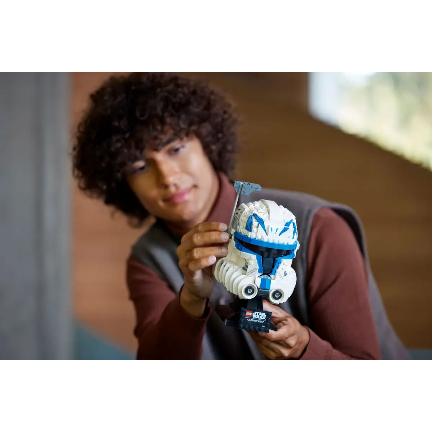 A person holding a Lego Star Wars Captain Rex helmet. The helmet is white and blue. The person has curly hair and is smiling.