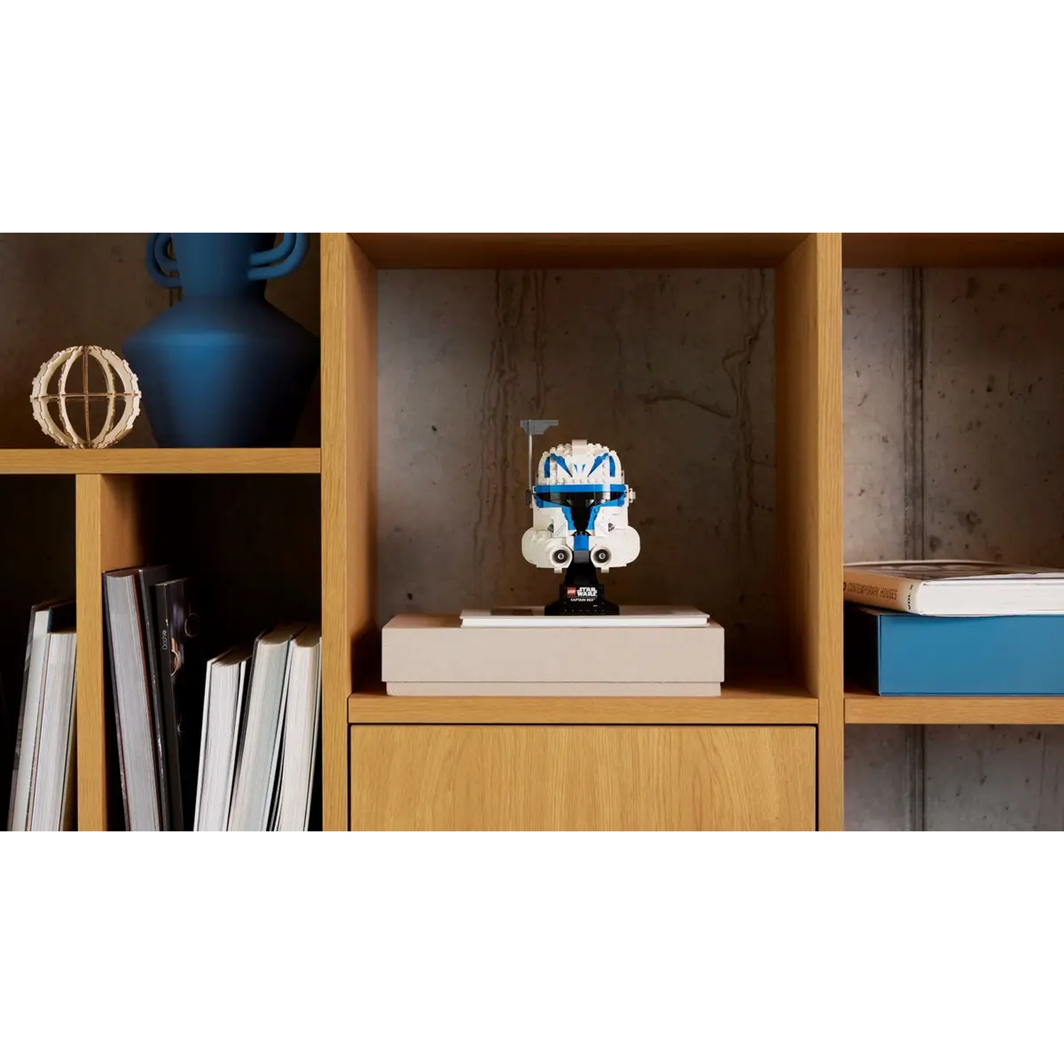 A Lego Star Wars helmet sits on a shelf. A blue vase, a wood sphere and books are on the shelf. The shelf is wood.