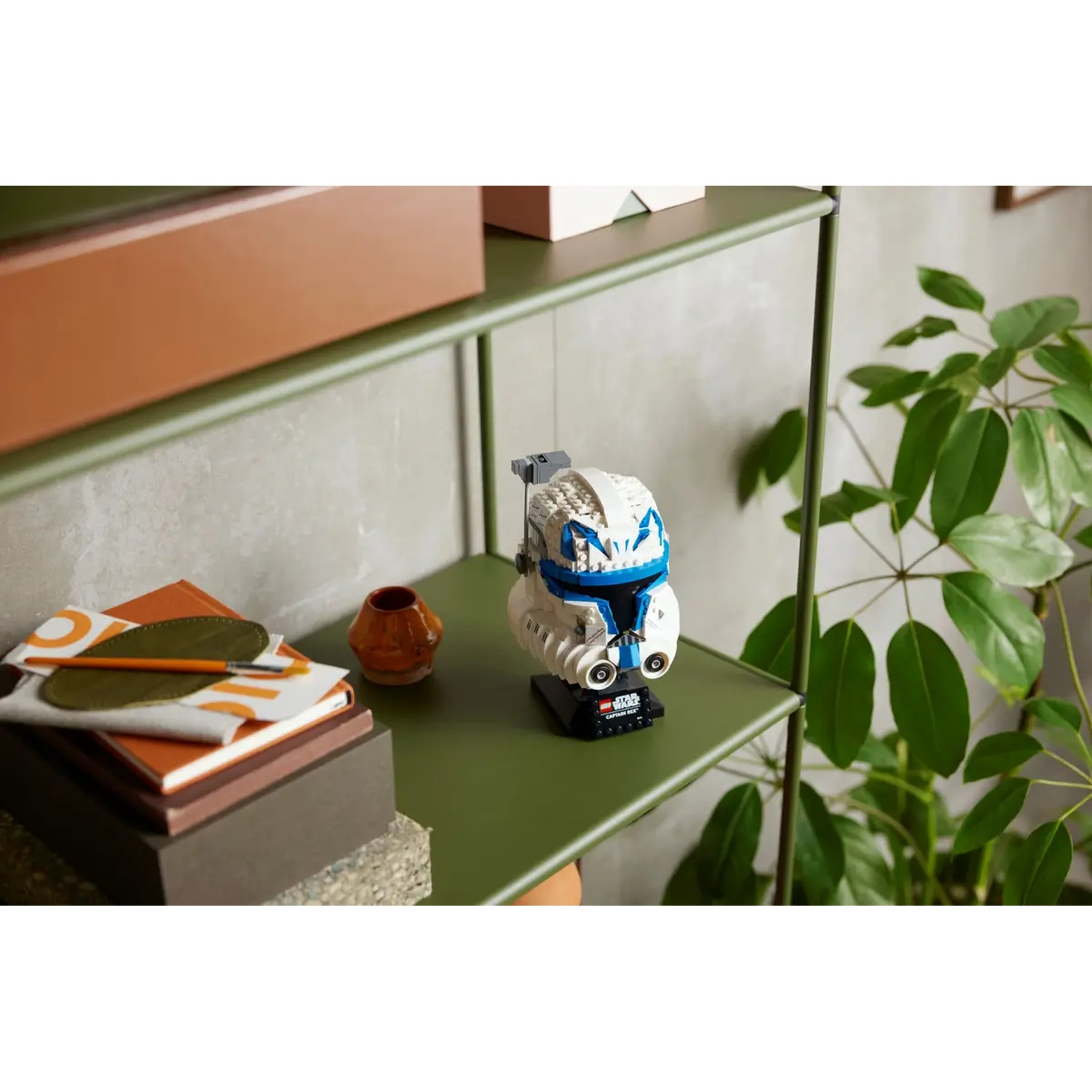 A Lego Star Wars Captain Rex helmet is on a green shelf, with books, a vase, and a plant nearby.