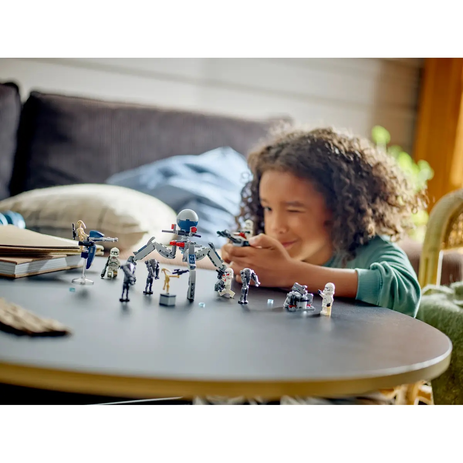 Een kind knipoogt naar de camera en speelt met Star Wars Lego op een tafel. Verschillende minifiguren en een spinnendroid zijn zichtbaar.