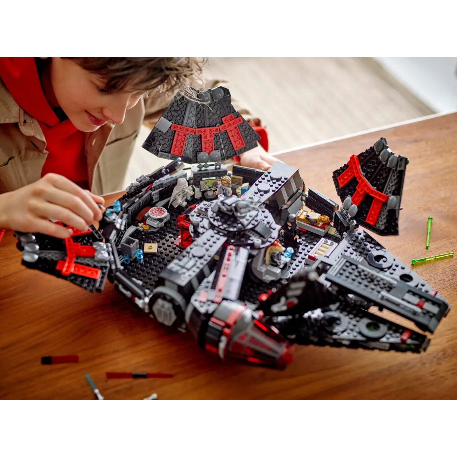 A person is building a black and red Lego spaceship on a wooden table. The spaceship is partially assembled, with details visible.