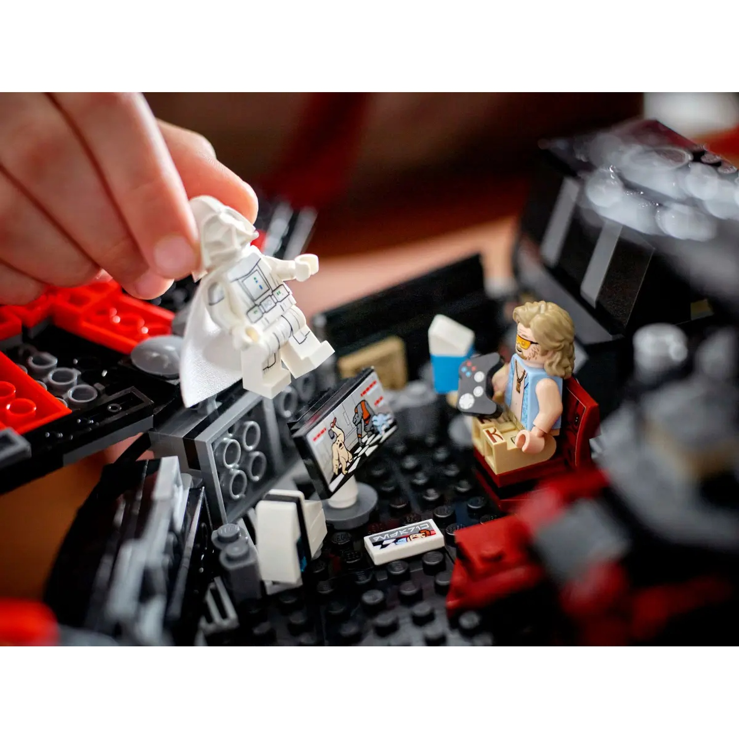 A hand holds a white Lego figure. A Lego figure with sunglasses is sitting. Lego blocks in red, black, and gray are present.