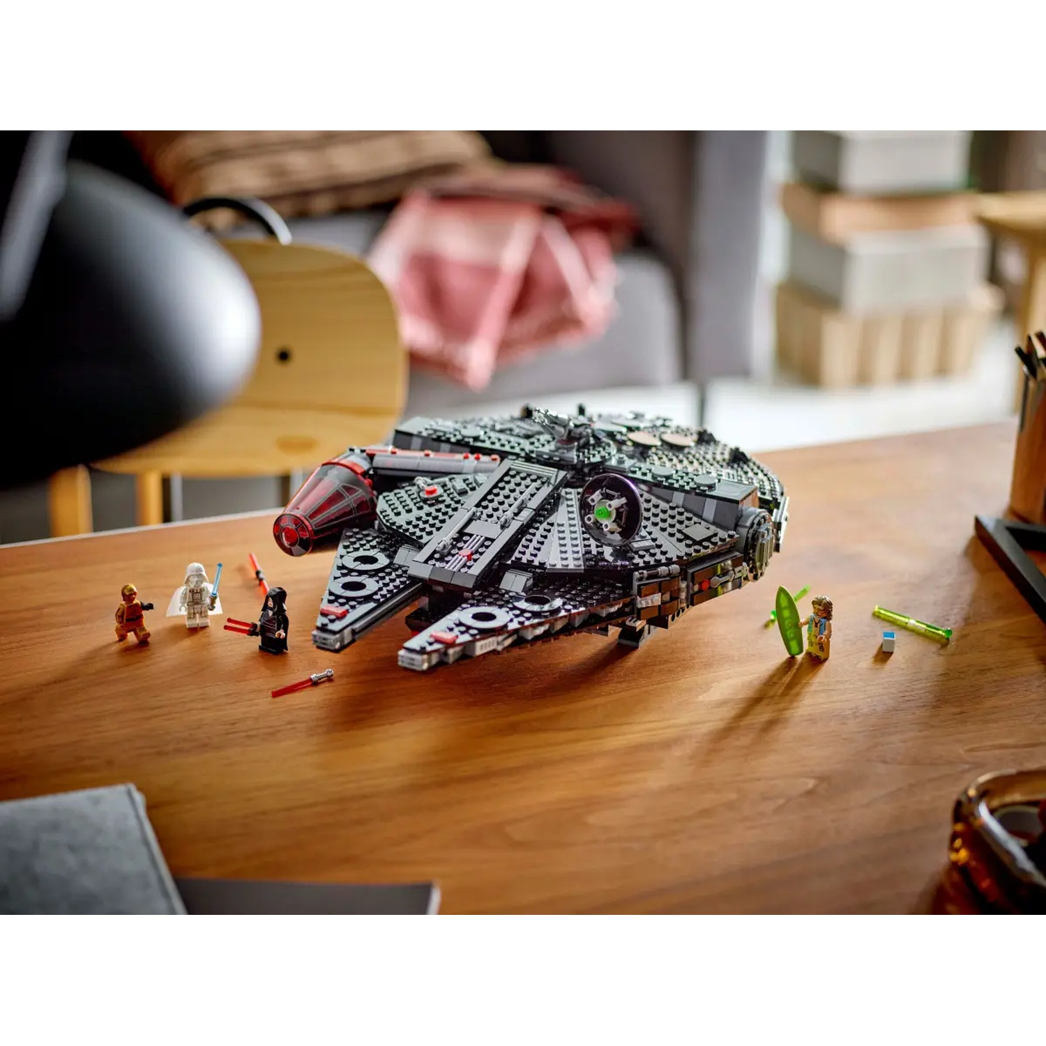 A Lego Millennium Falcon model with mini-figures on a wooden table. Background includes a chair and a grey sofa.