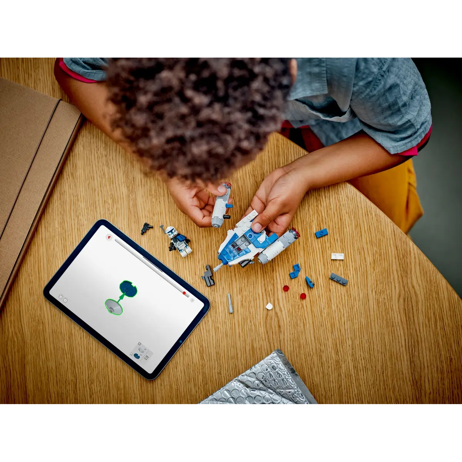 A person builds a Lego Star Wars ship on a wooden table. A tablet shows instructions. Various Lego pieces are scattered around.
