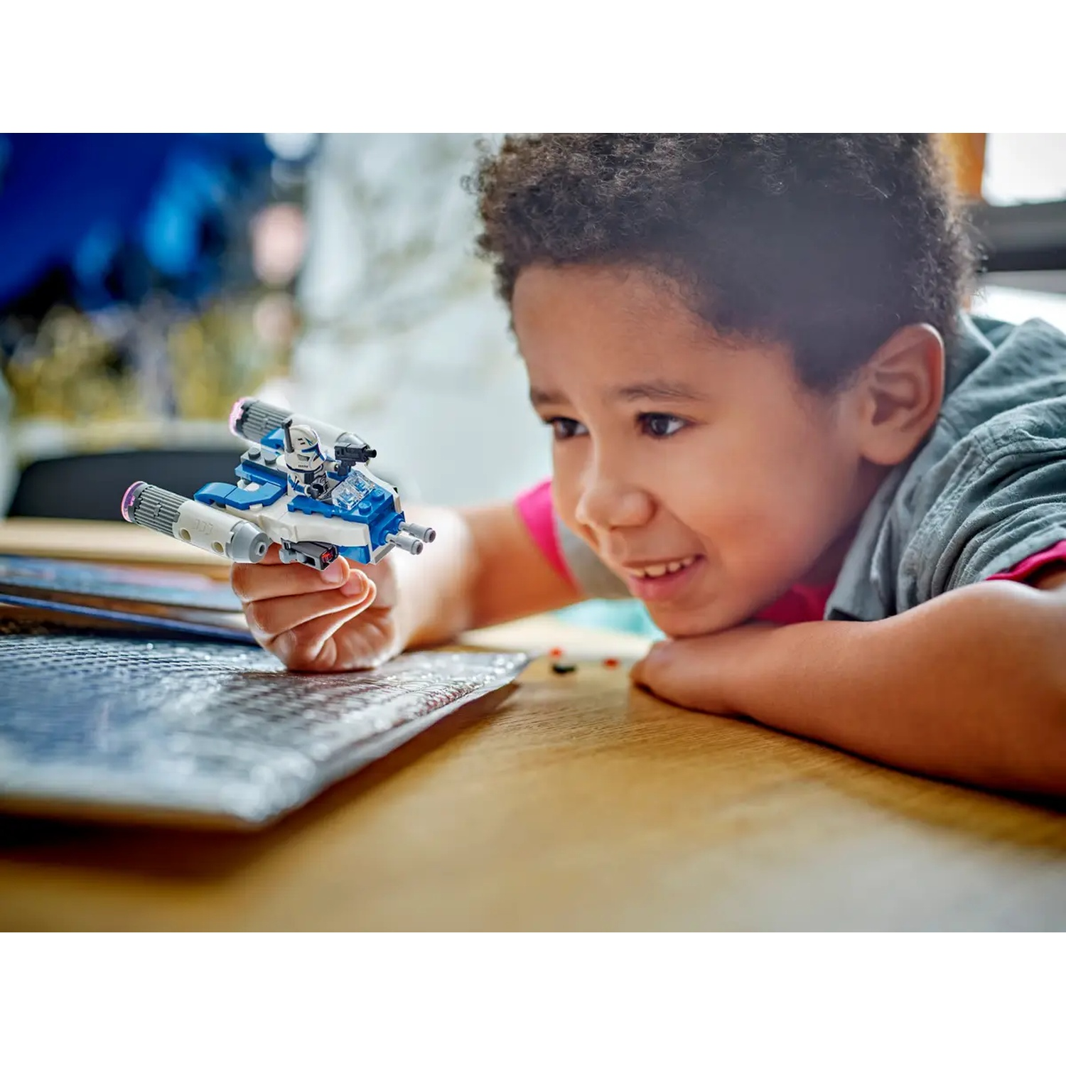 A smiling boy looks at a Lego starfighter he holds. It's white and blue. He's at a wooden table, and a silver mat is visible.