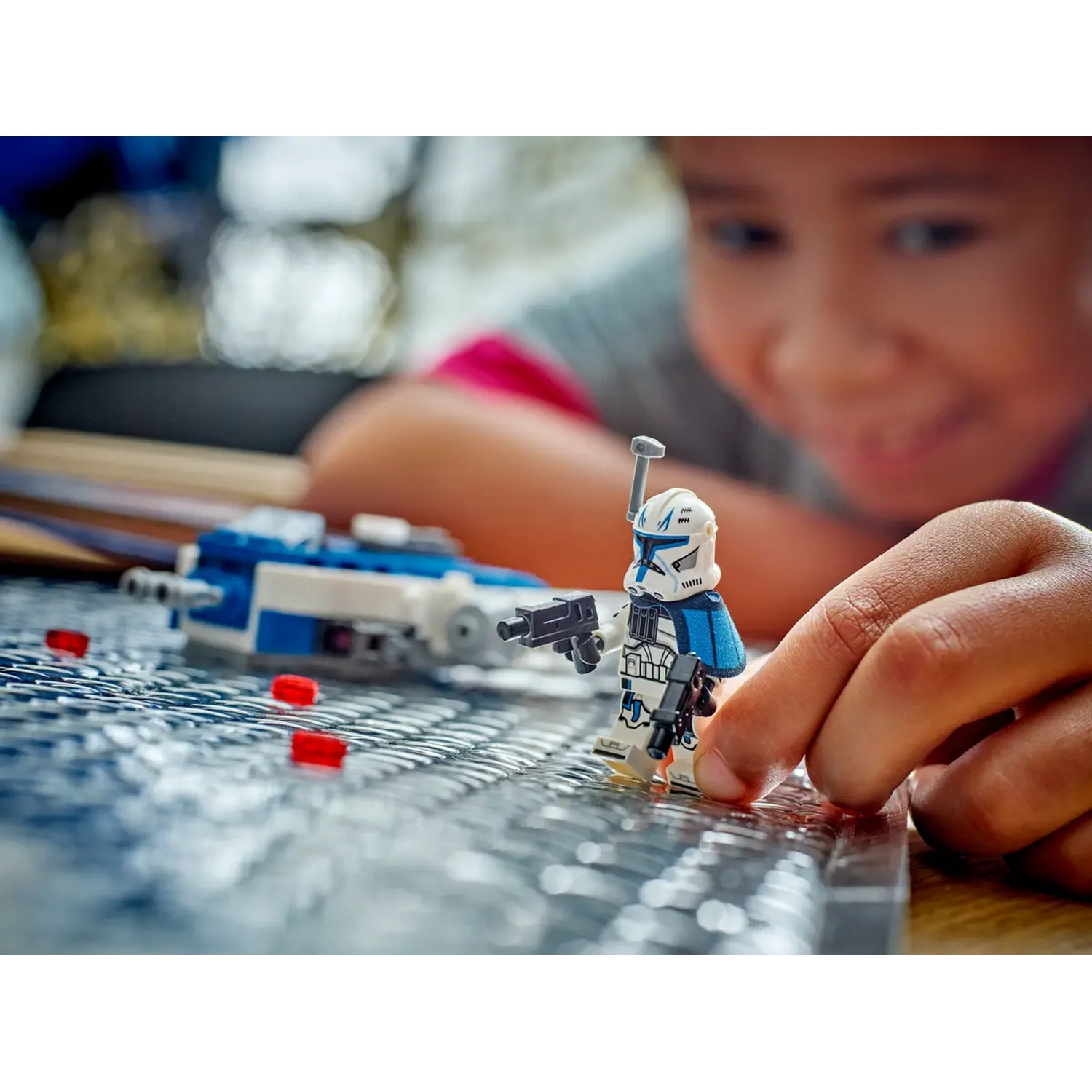 A child's hand holding a Lego figurine. A blue and white Lego ship and red blocks are in the background.