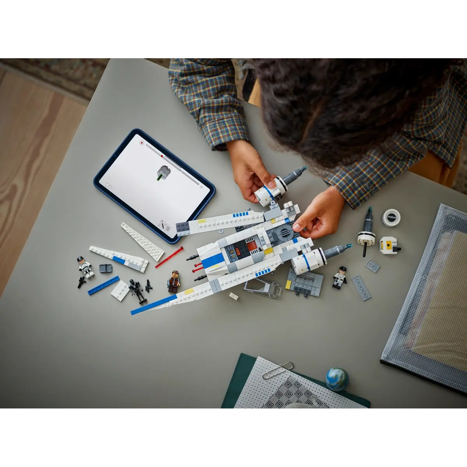 A person assembling a Lego Star Wars X-wing fighter on a gray table. Includes tablet, mini figures, and various Lego pieces.