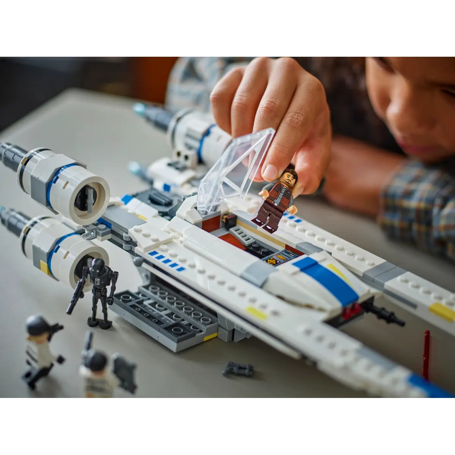 A child's hand places a Lego figure into a white and blue X-wing fighter. Other Lego figures and parts are nearby.