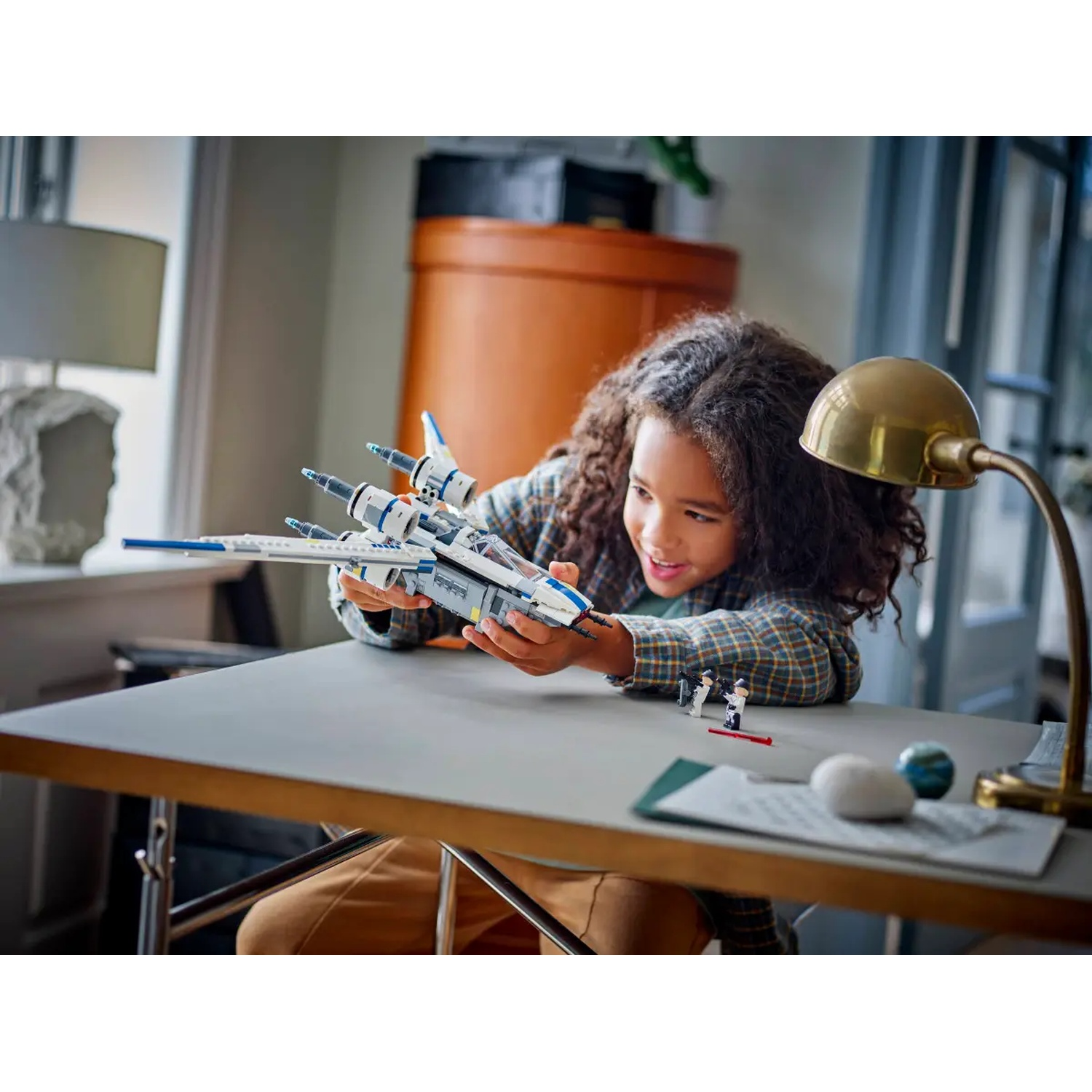 A girl holds a toy spaceship. There are small figurines and a lamp on the table. The girl is wearing a plaid shirt.