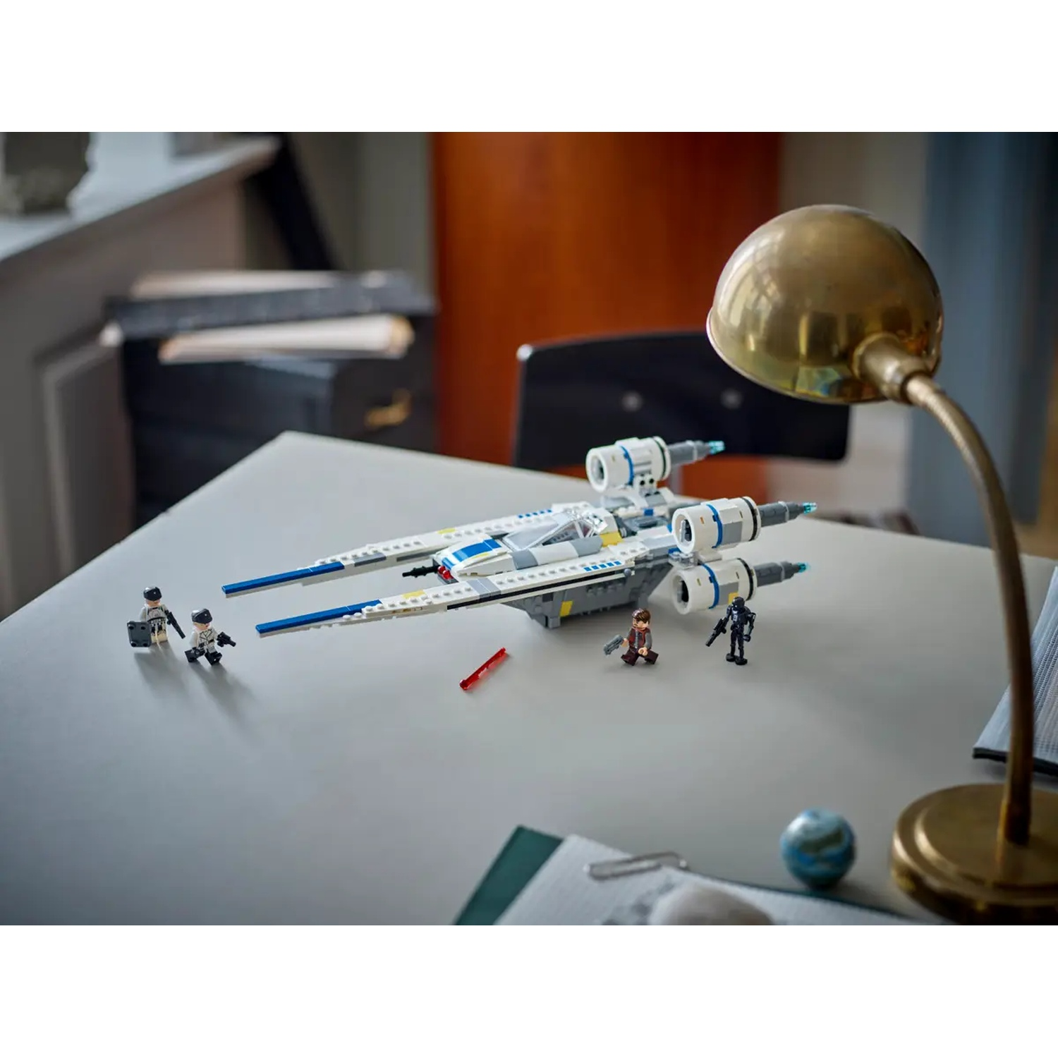A white and blue Lego spaceship sits on a table with Lego figures. A lamp is to the right. A small globe and papers are also present.