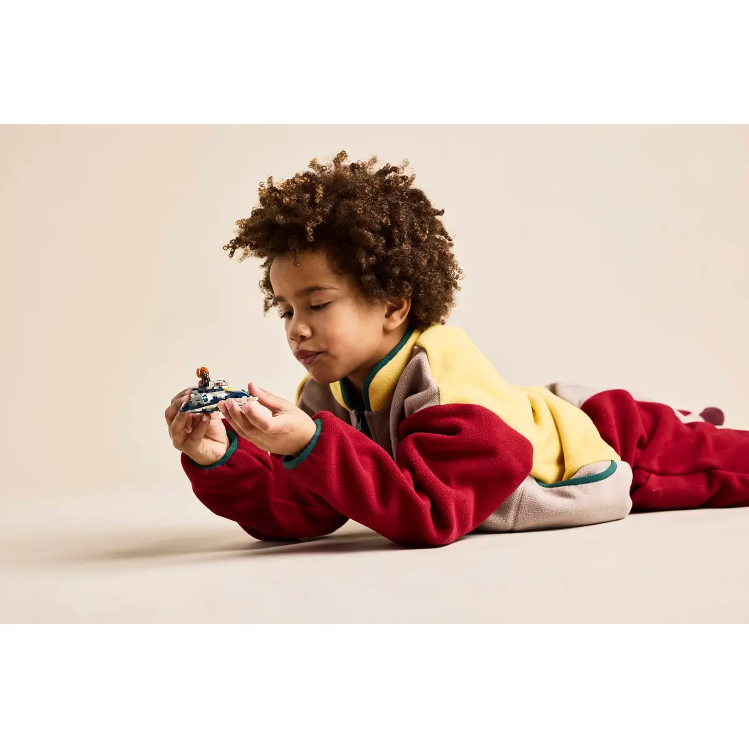 A child in a colorful outfit lies on the floor, holding a small toy car. The background is a plain beige color.