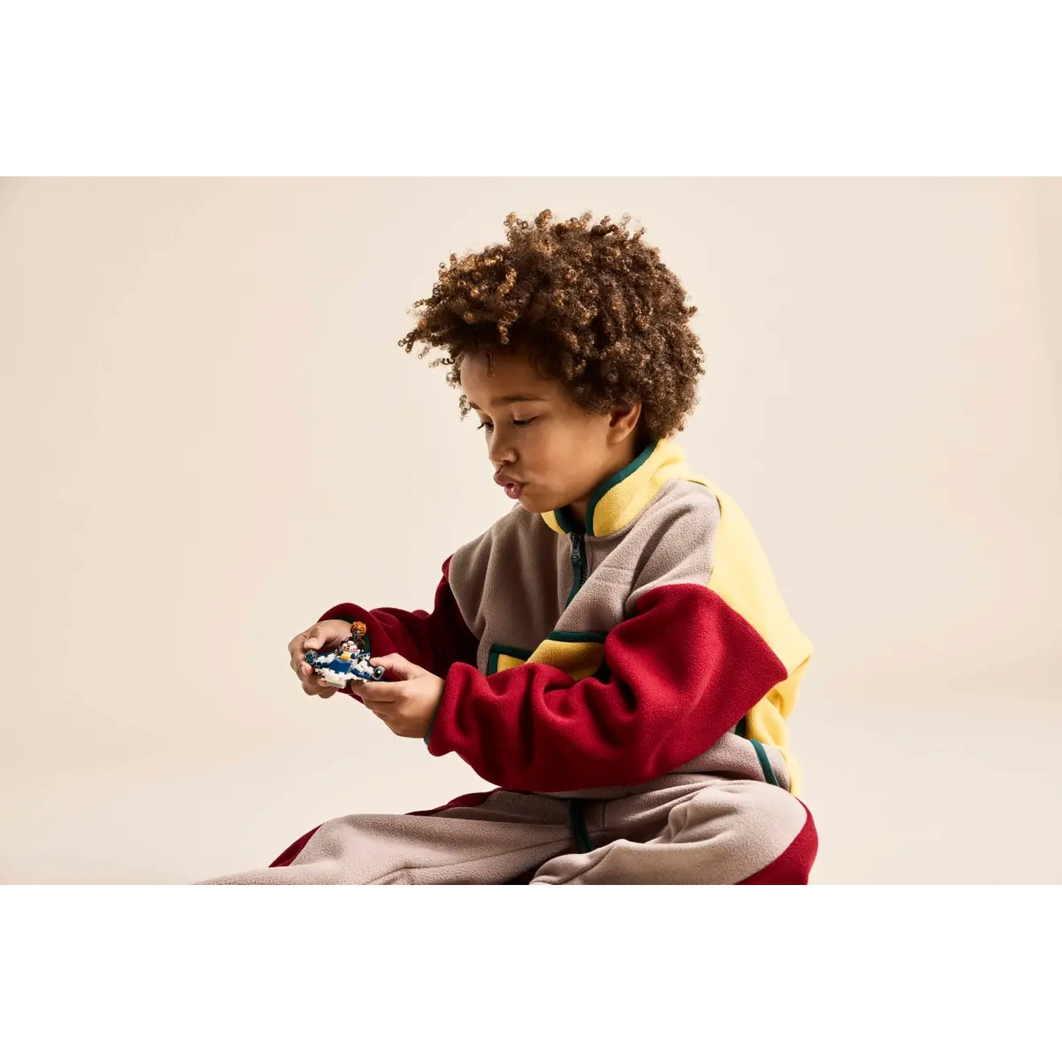 A child in a color-block outfit holds a small toy. The outfit is yellow, red, and gray. The background is cream-colored.