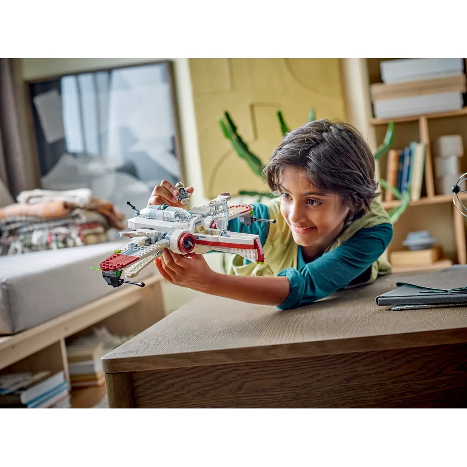 A boy holds a toy X-wing fighter. He smiles. The background shows a room with a window, bookshelves, and a desk.