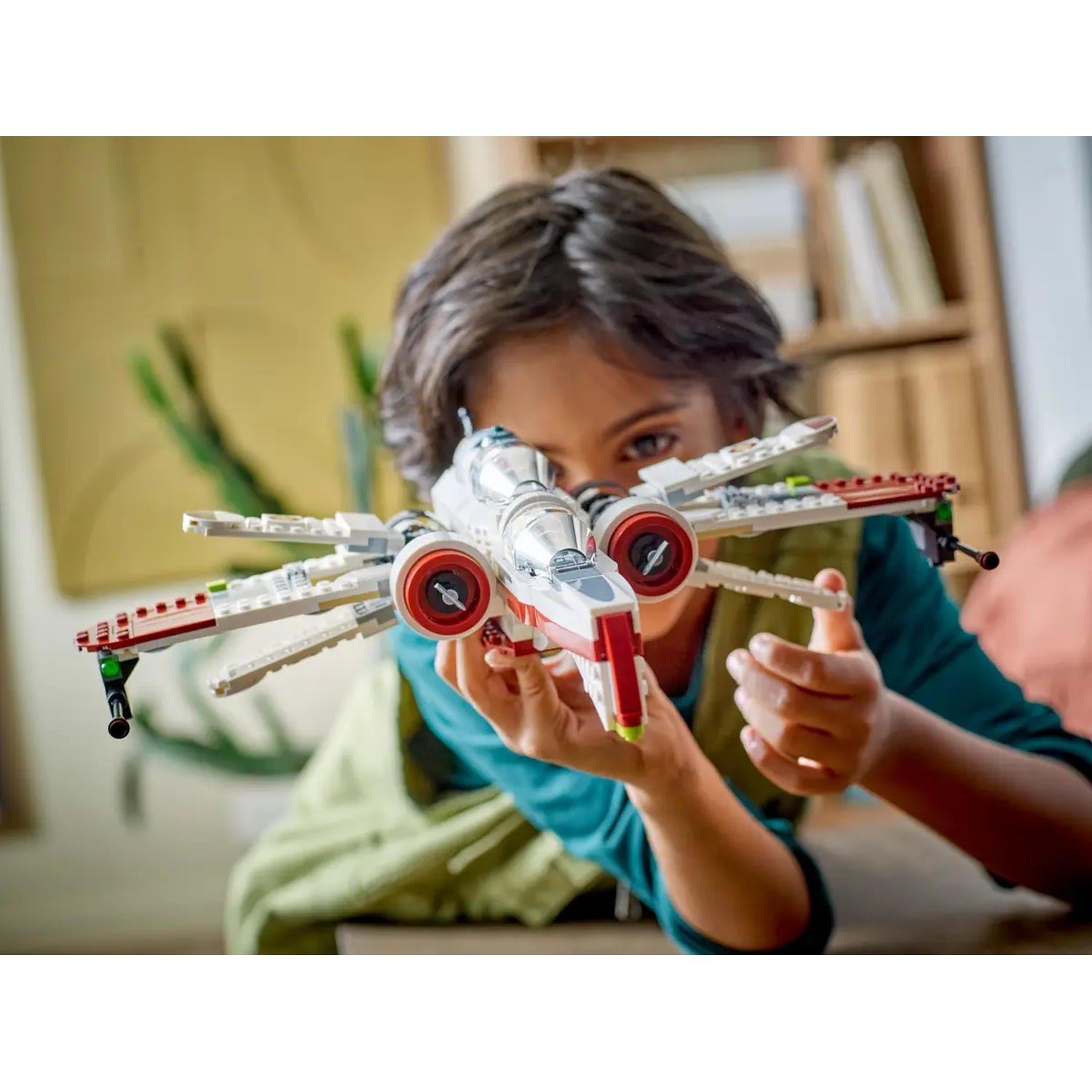 A child holds a LEGO Star Wars X-wing fighter. It's white and red, with the child's face partially visible. Background shows a room.