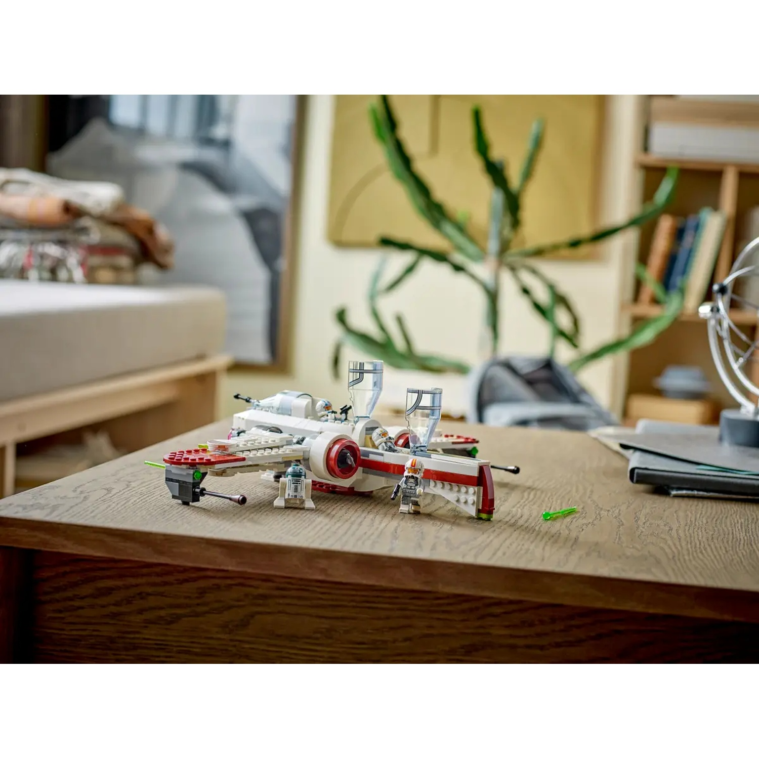 A Lego X-wing fighter is on a wooden table. Figures and other Lego pieces are nearby. A plant and bookcase are in the background.