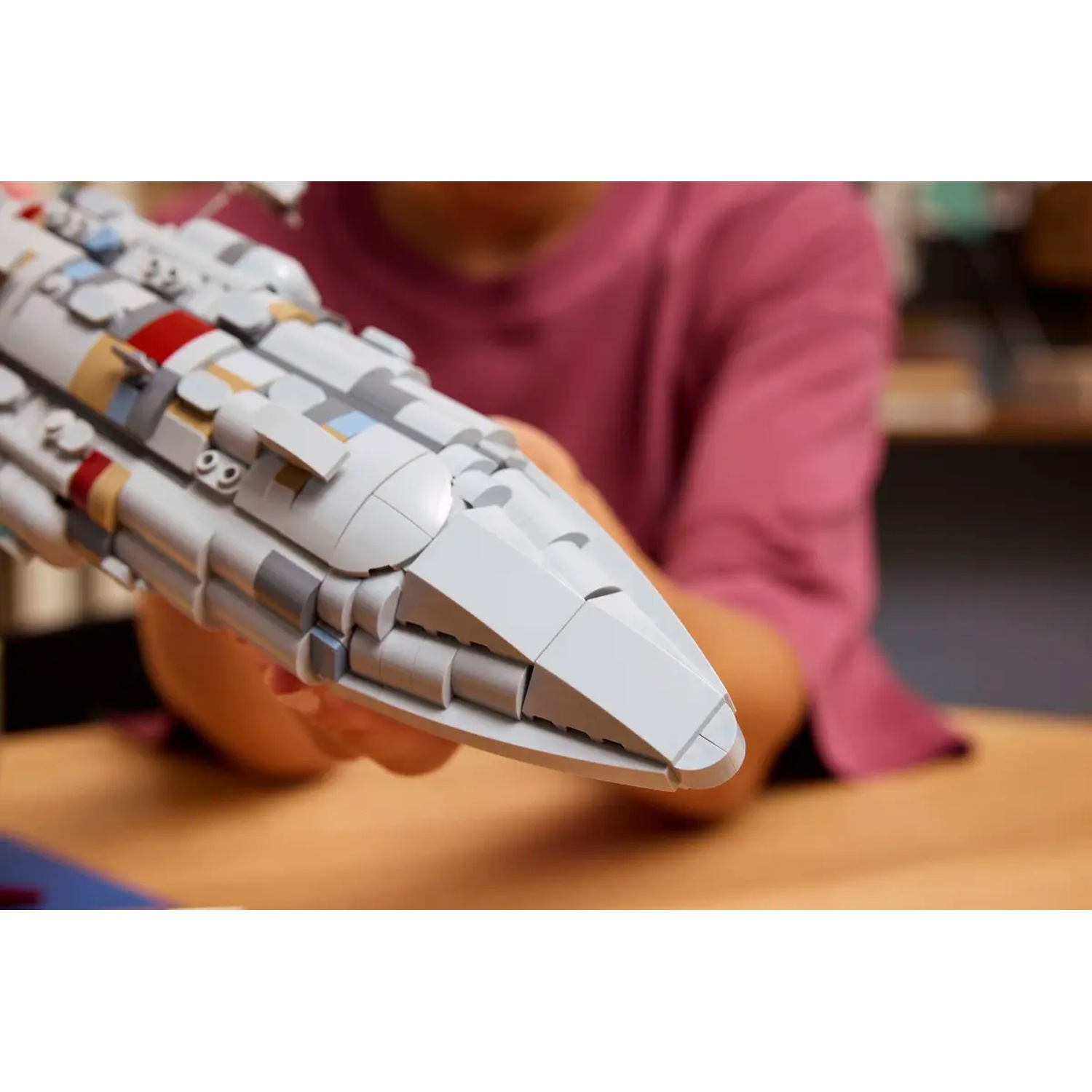 A person holds a gray Lego spaceship. The ship is angled toward the viewer. The background is a pink shirt and wooden table.