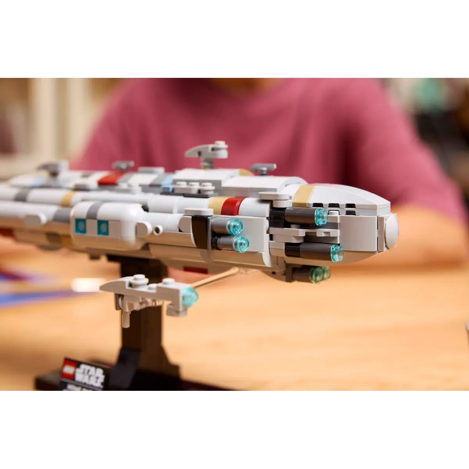 A Lego Star Wars model, mostly white and gray, on a black stand. A person in a pink shirt is blurred in the background.