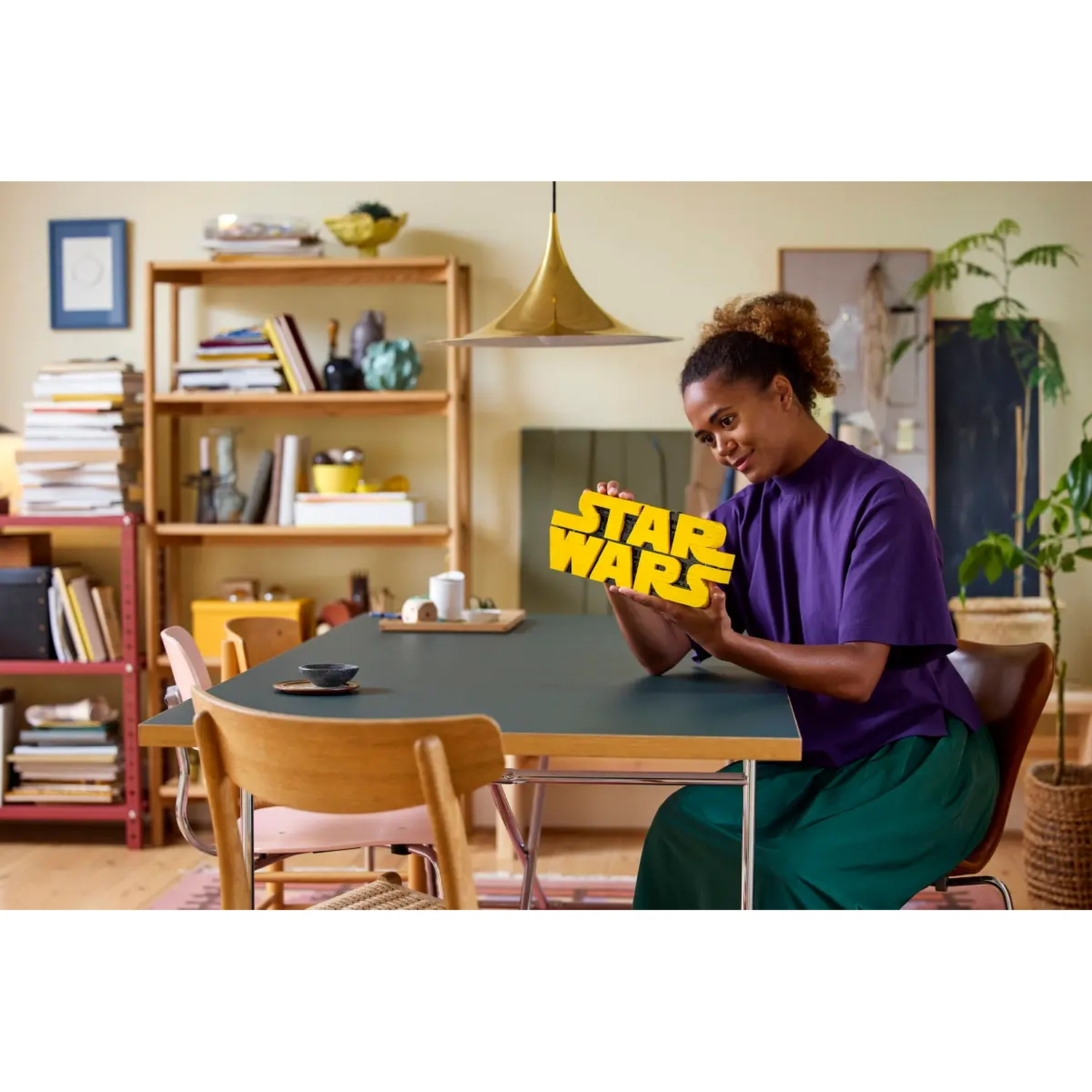 Een vrouw houdt een geel 'STAR WARS' bord vast. Ze zit aan een tafel. Boekenplanken en planten op de achtergrond.