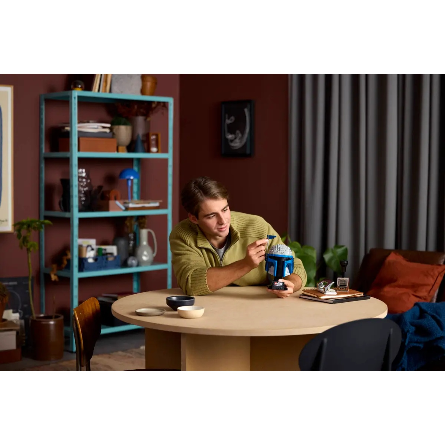 A man in a green sweater is holding a blue and gray helmet at a table. Bowls and other items are on the table.