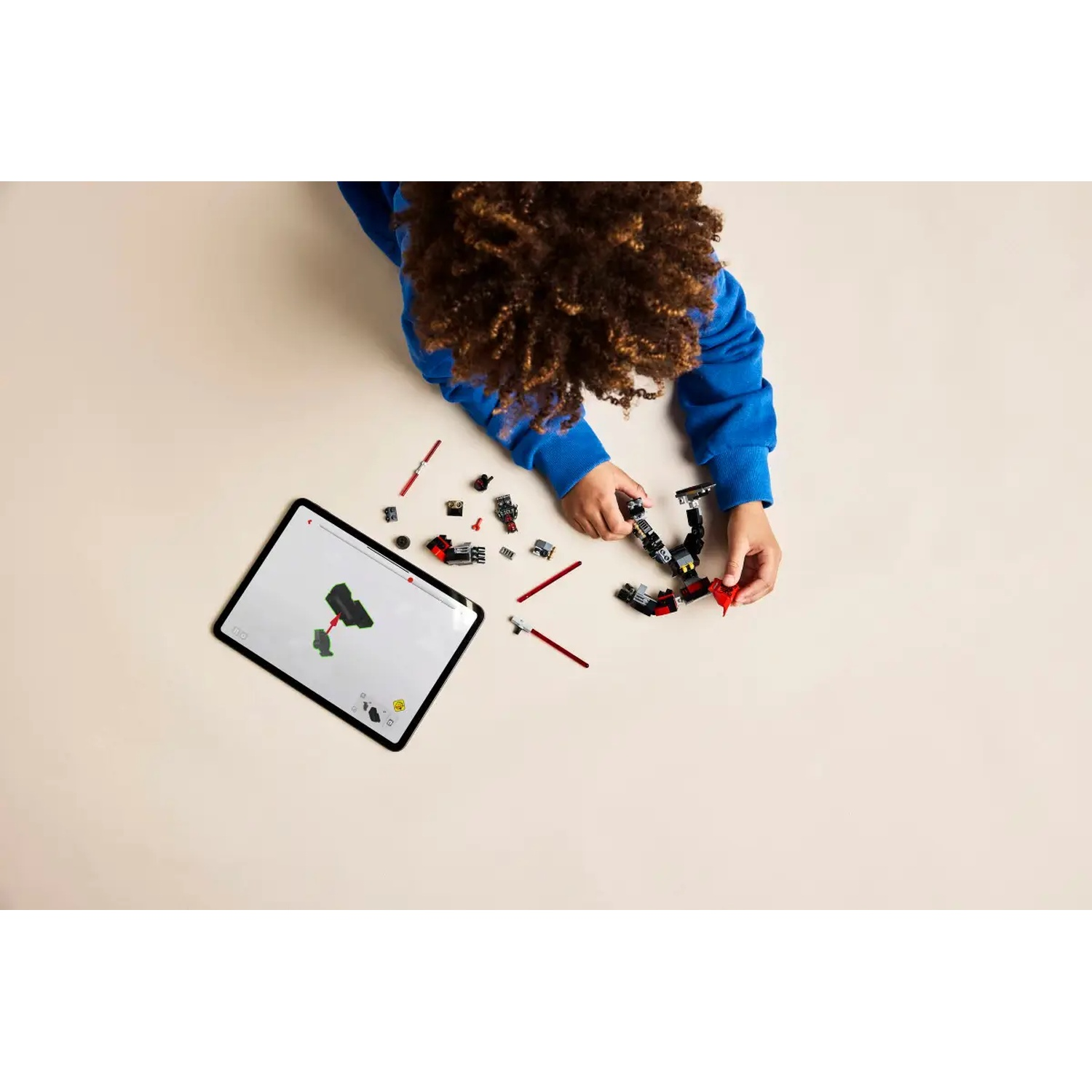 Een kind in het blauw speelt met Lego, instructies volgend op een tablet. Rode en zwarte Lego-stukken liggen verspreid.