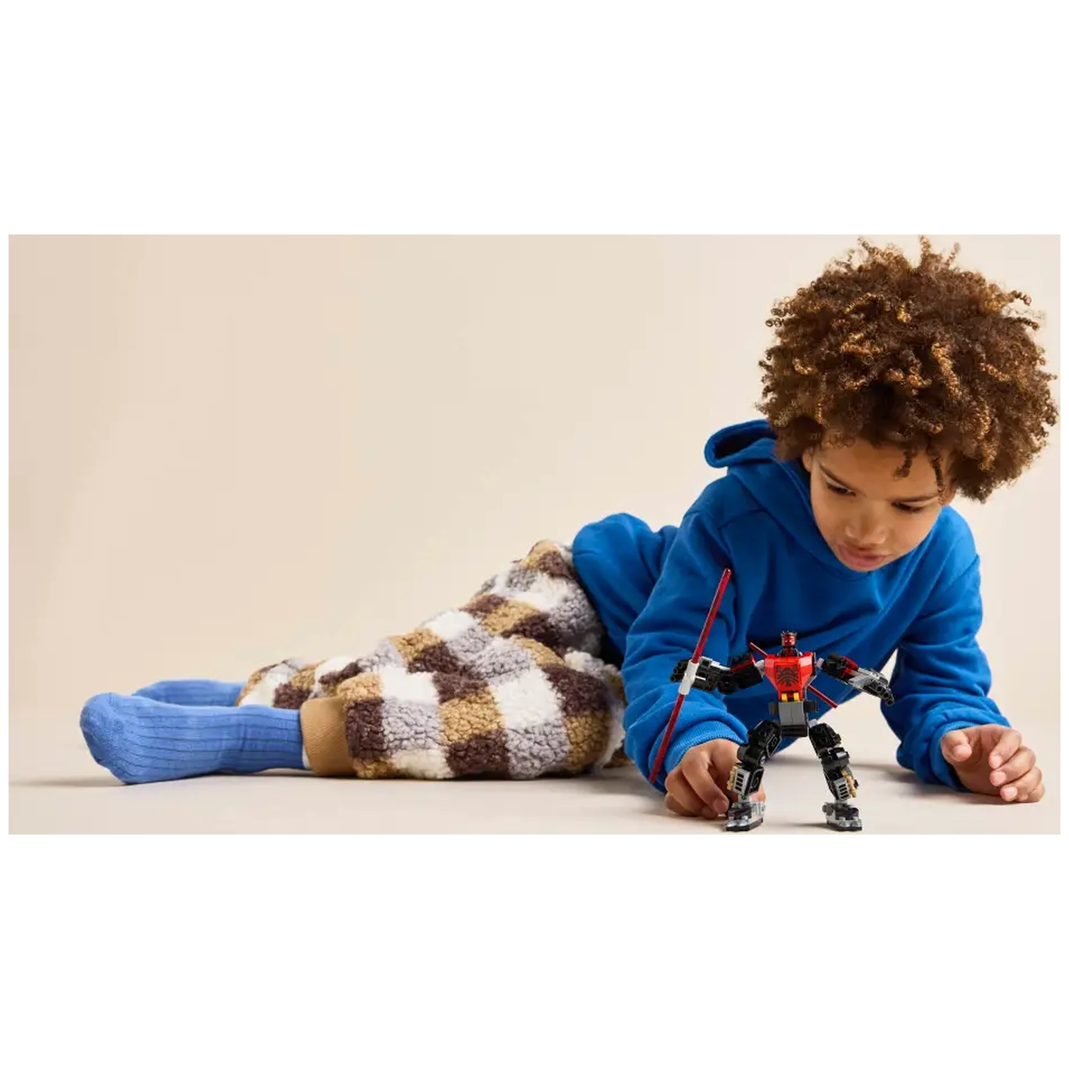 Een kind in een blauwe hoodie en een broek met patroon speelt met een rood-zwarte robotspeelgoed op een lichte achtergrond.
