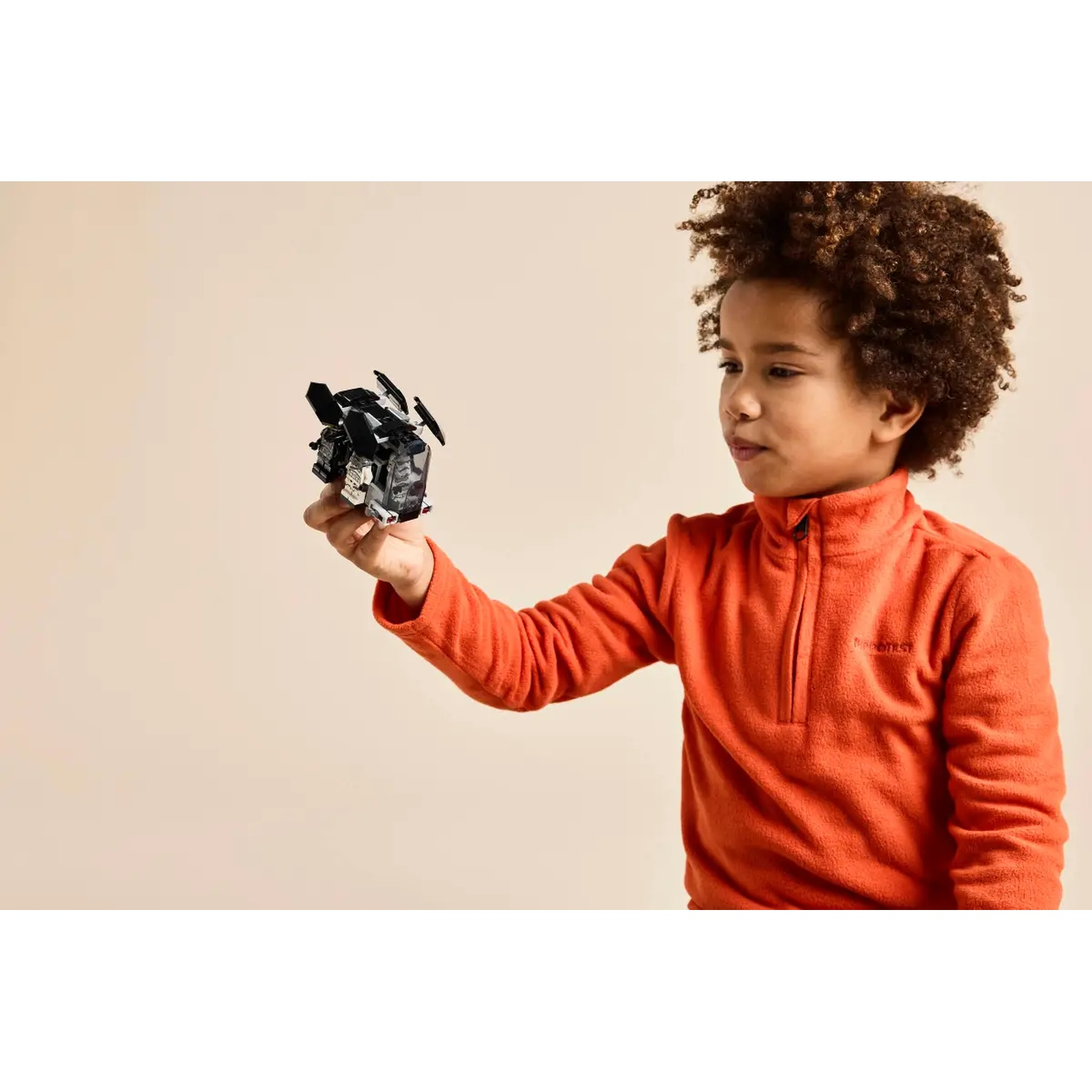 A young boy in an orange sweater holds a black toy airplane against a beige background.