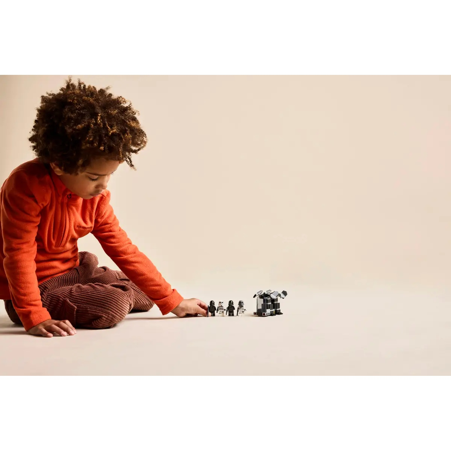 A child in orange looks at Lego figurines and a building. The child is wearing an orange jacket and brown pants, on a beige background.