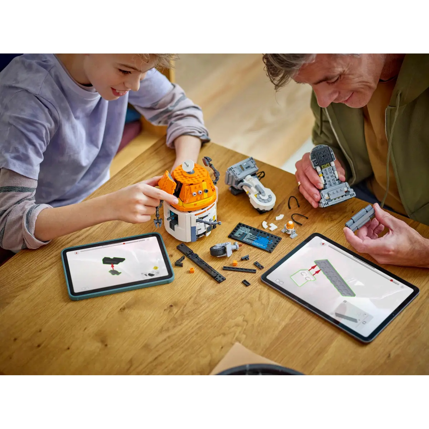 Two people building Lego, using tablets for instructions. Orange and white Lego structure is in progress. Parts are scattered on a wooden table.