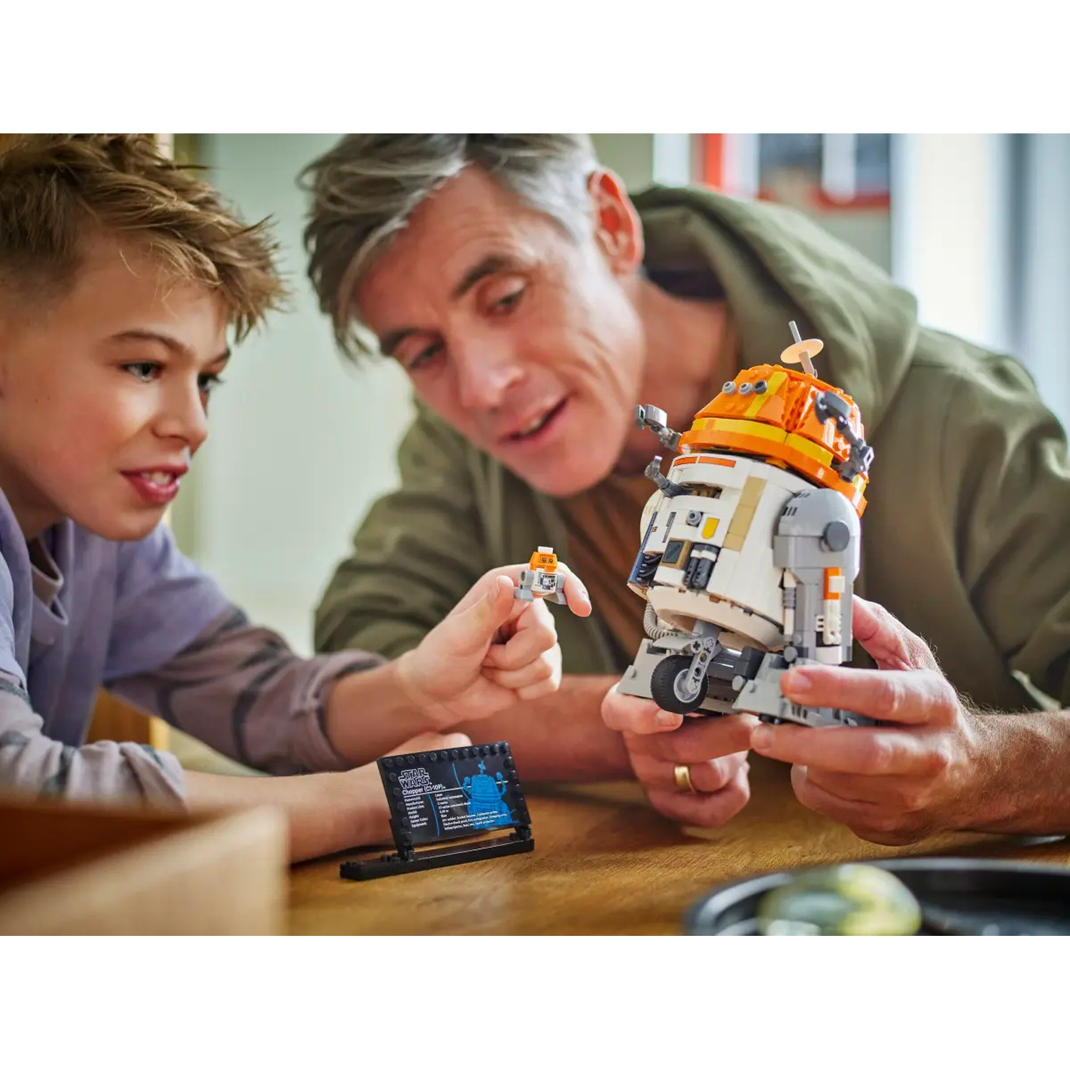 A man and a boy are assembling a Lego Star Wars droid. The droid is white, orange, and gray. A small figure is held.