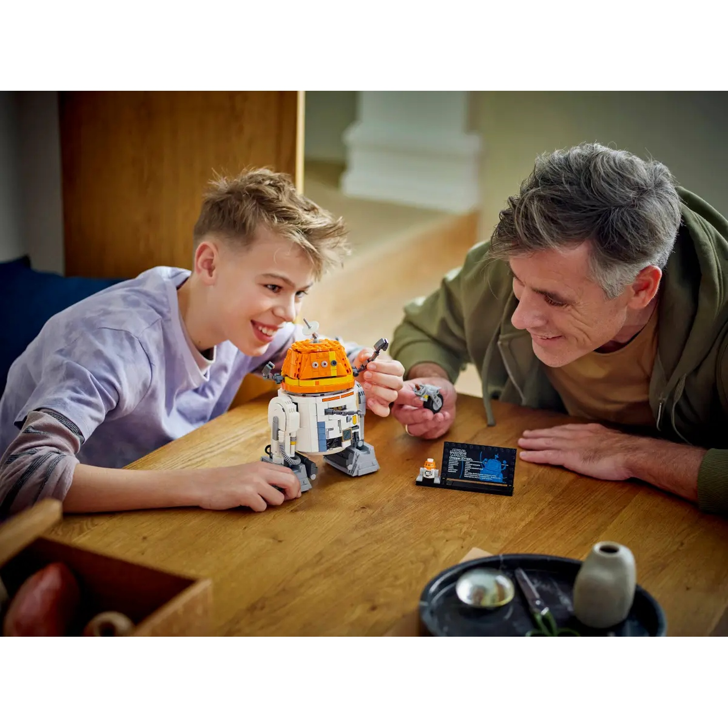 A father and son are building a Lego Star Wars droid together at a wooden table. They are smiling and looking at the droid.