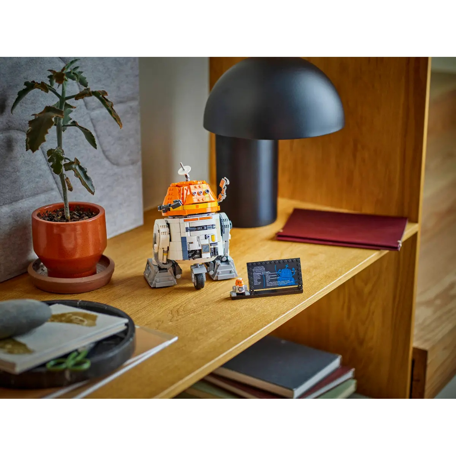 A Lego droid with orange and white parts sits on a wooden shelf. A plant in a terracotta pot and a black lamp are also on the shelf.