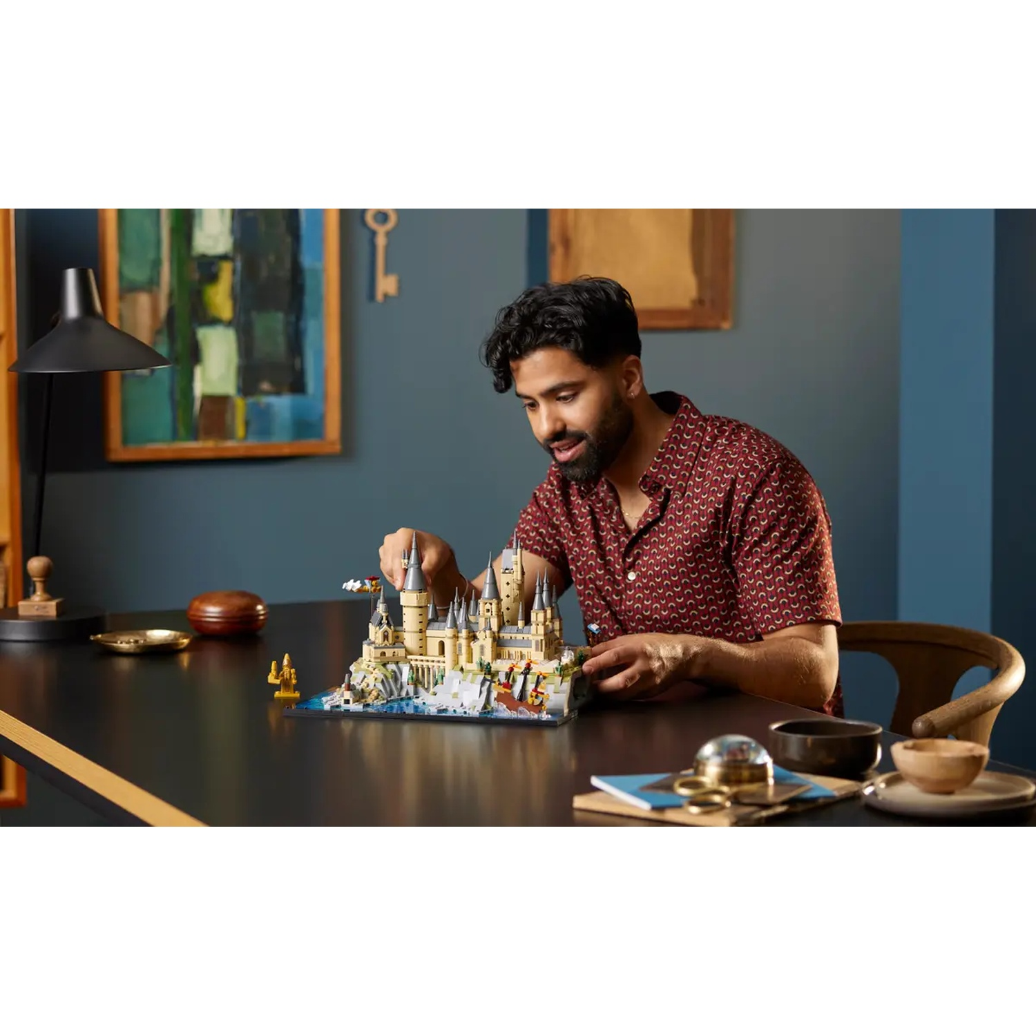 Een man bouwt een Lego Hogwarts-model op een donkere tafel, met diverse items eromheen. Achtergrond is kunst en een lamp.
