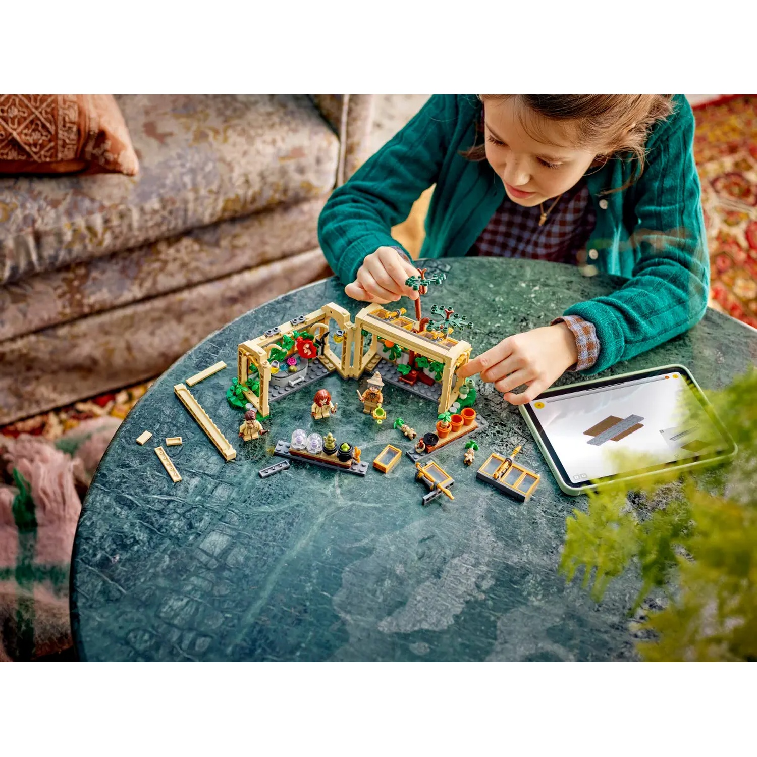 A girl is playing with a Lego set on a green table. The set depicts a greenhouse, and a tablet is visible to the right.