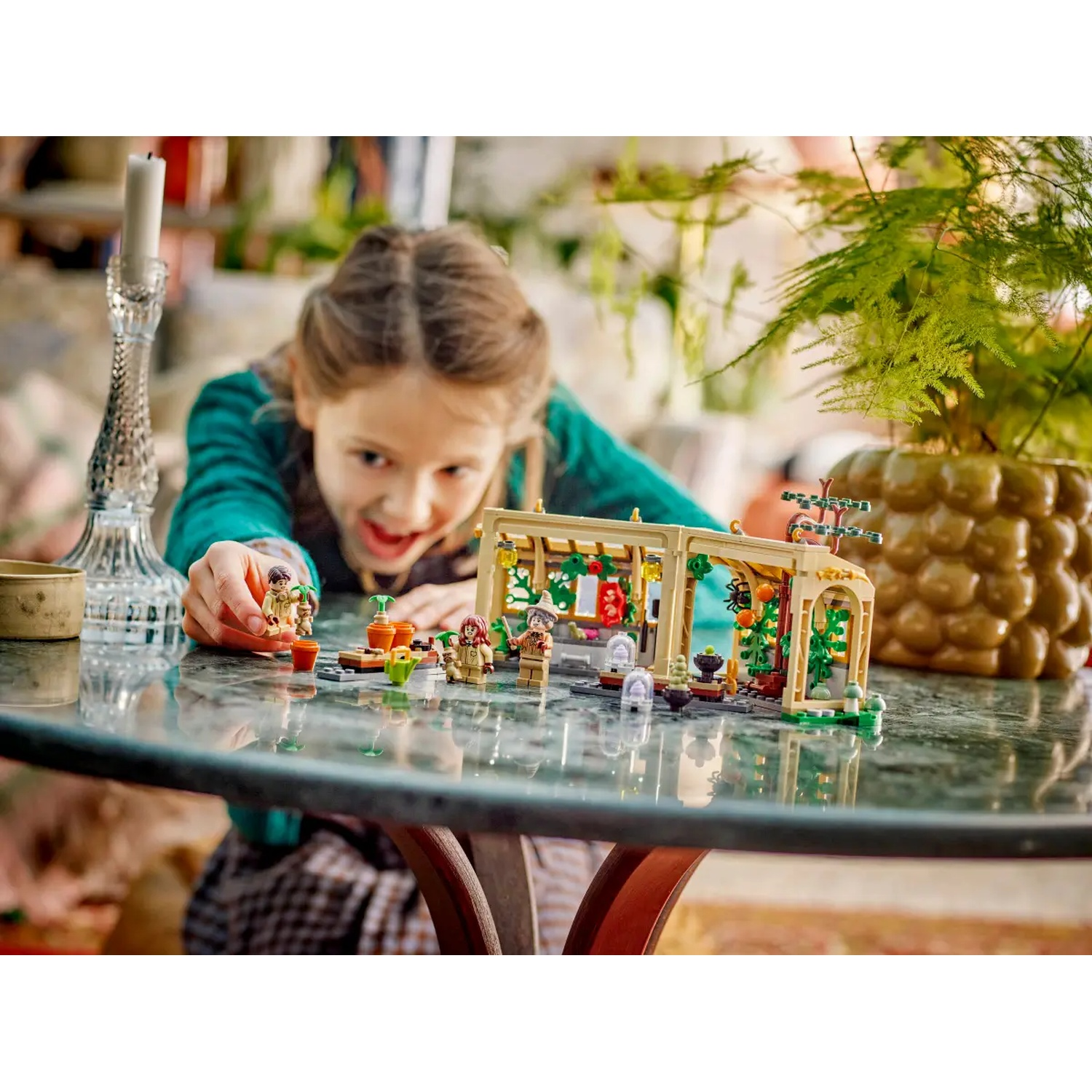A girl plays with a Lego set on a glass table. The set includes figures and a greenhouse structure.