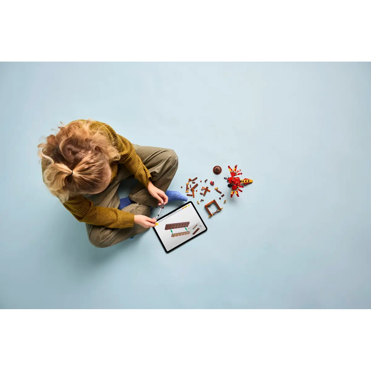 Een kind zit en bekijkt een tablet met bouwinstructies. Legostenen en een voltooid model liggen in de buurt.