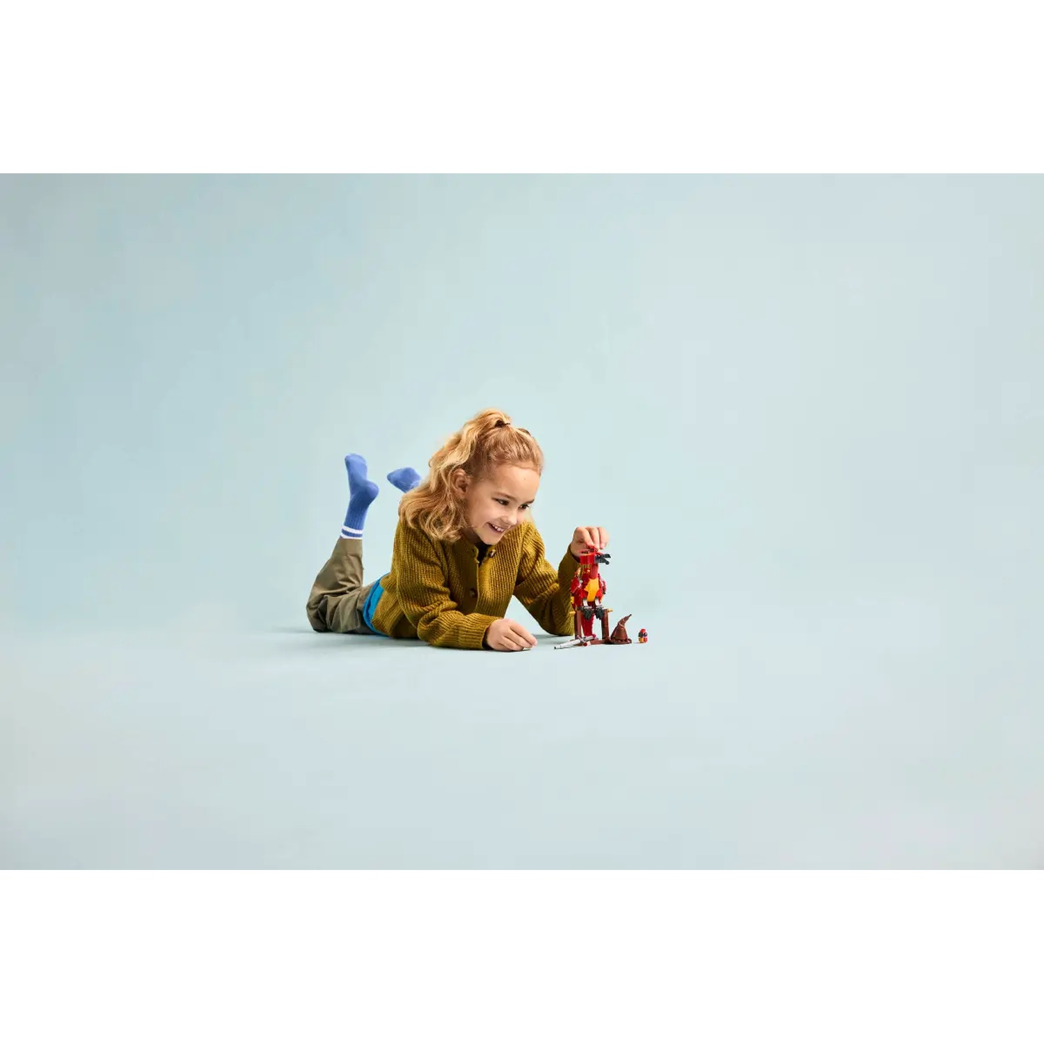 Een meisje ligt op de grond en speelt met een Lego-set. Ze draagt een gele trui en blauwe sokken. De set is rood en bruin.