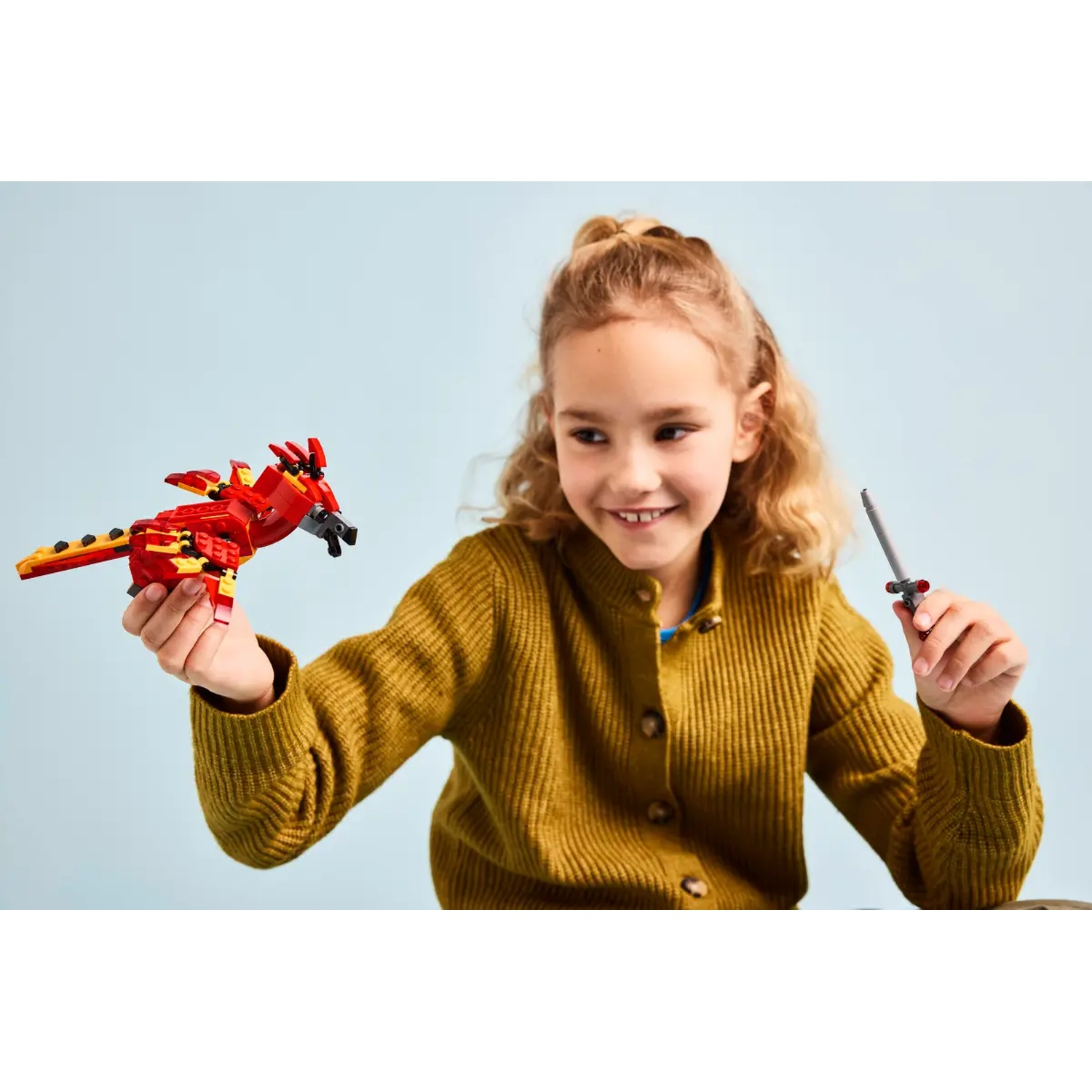 Een meisje glimlacht, met een rode Lego-draak en een grijs zwaard. Ze draagt een gele trui tegen een lichtblauwe achtergrond.