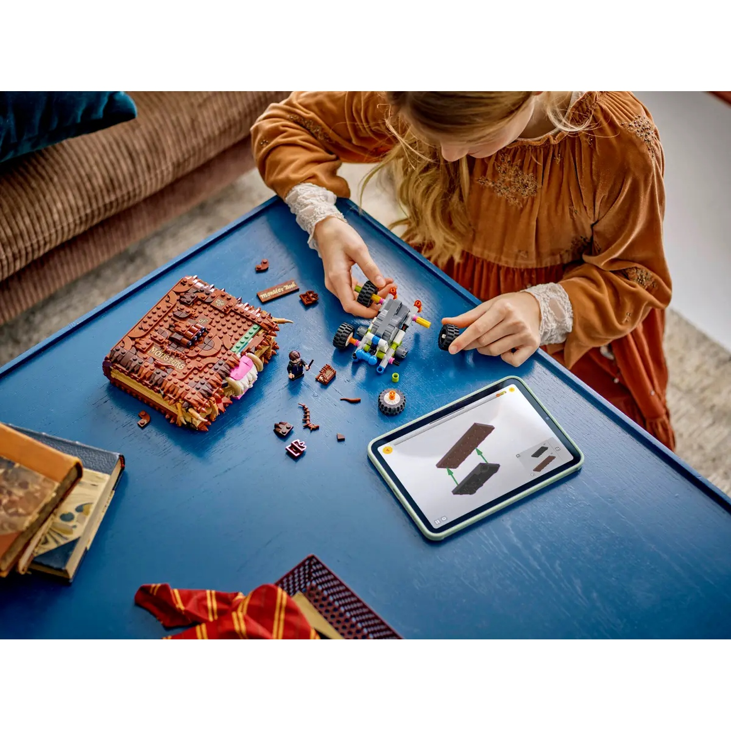 A girl assembles a Lego set on a blue table. Parts and a tablet showing instructions are present. A book and scarf are nearby.