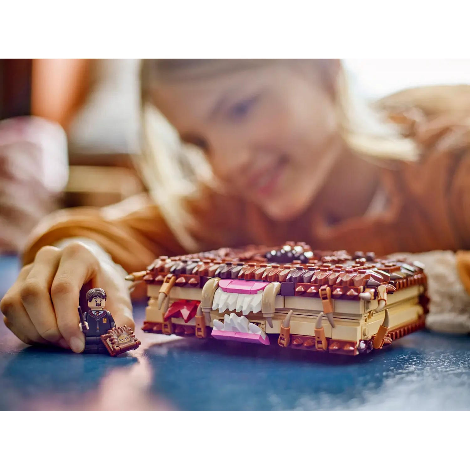 A child smiles, Lego book and figure in focus. The book is brown, teeth-like structures, a Lego figure stands in front.