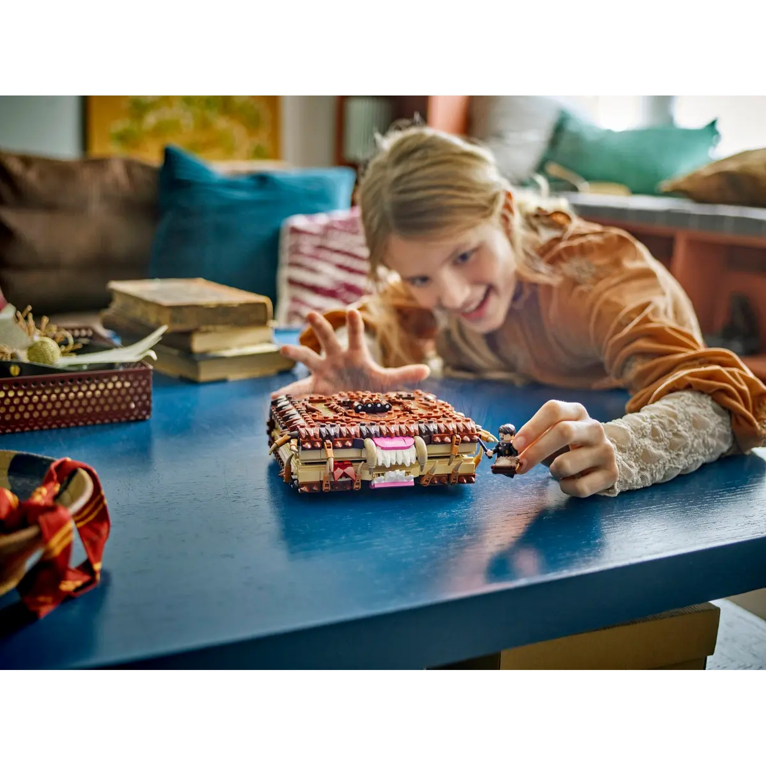 A girl smiles at a Lego book and figurine. The book is brown, pink, and tan on a blue table. Background includes pillows.