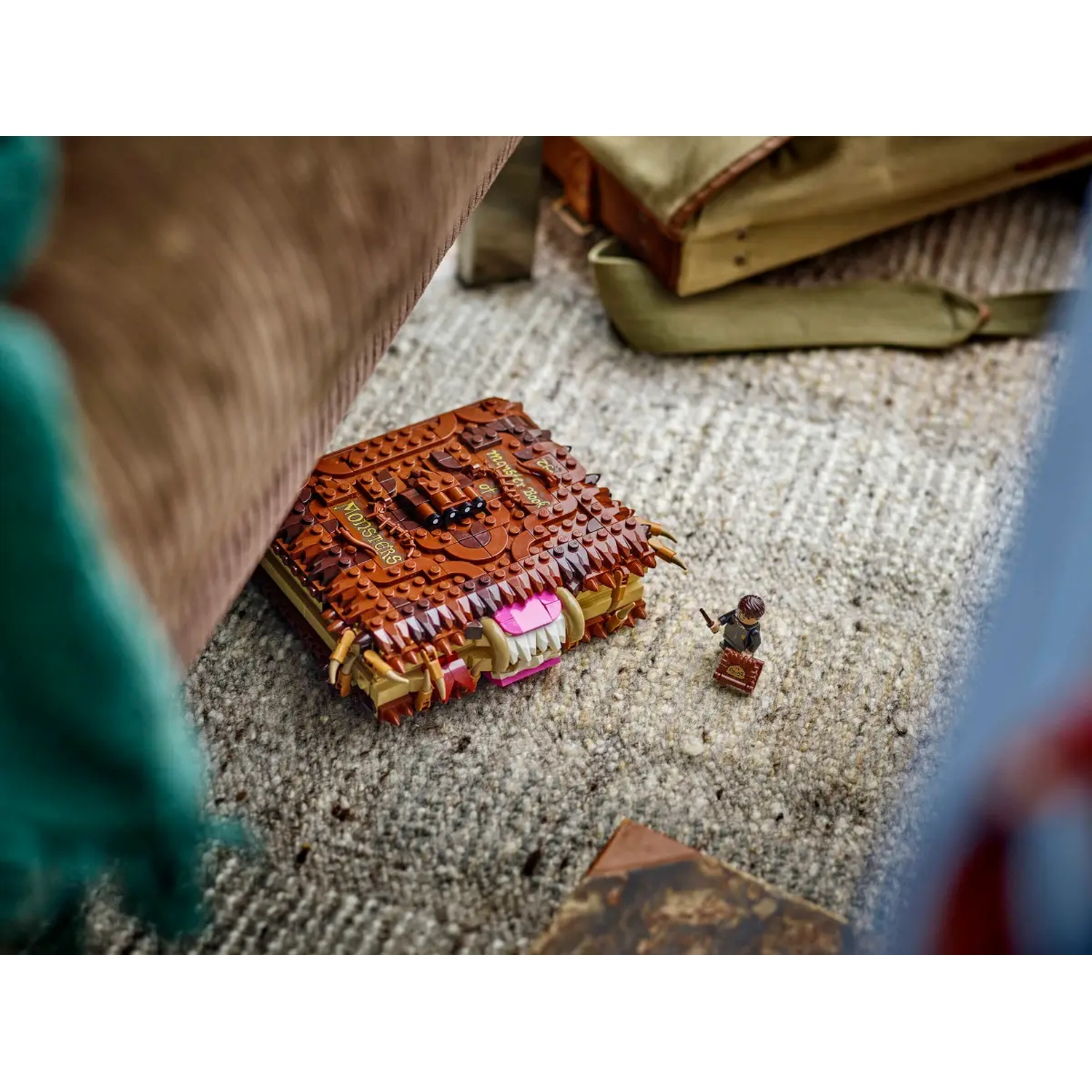 A Lego monster book with teeth sits on a rug. A Lego figure with a wand and a suitcase stands nearby. A bag is in the background.