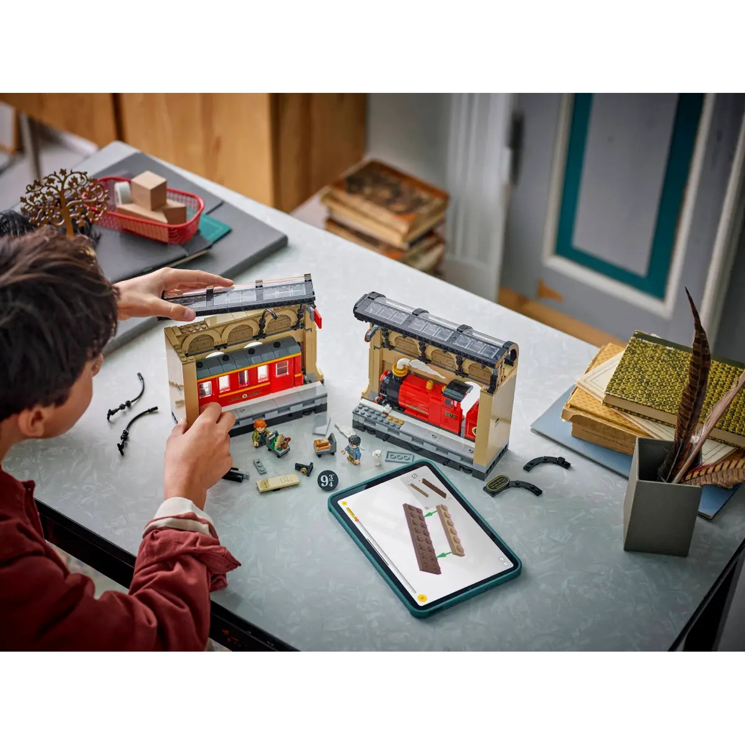 A child assembles a Lego train station. Red train cars are in stations. A tablet shows instructions. Other items are on the table.