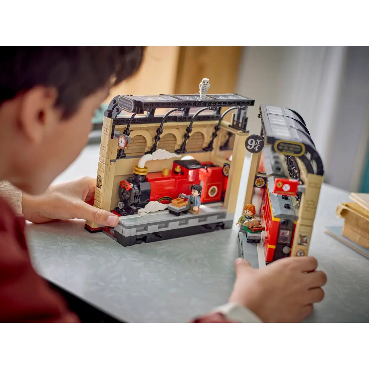 A child holds a LEGO Harry Potter set. It features a red train, platform 9 3/4, and minifigures. Background is a neutral table.