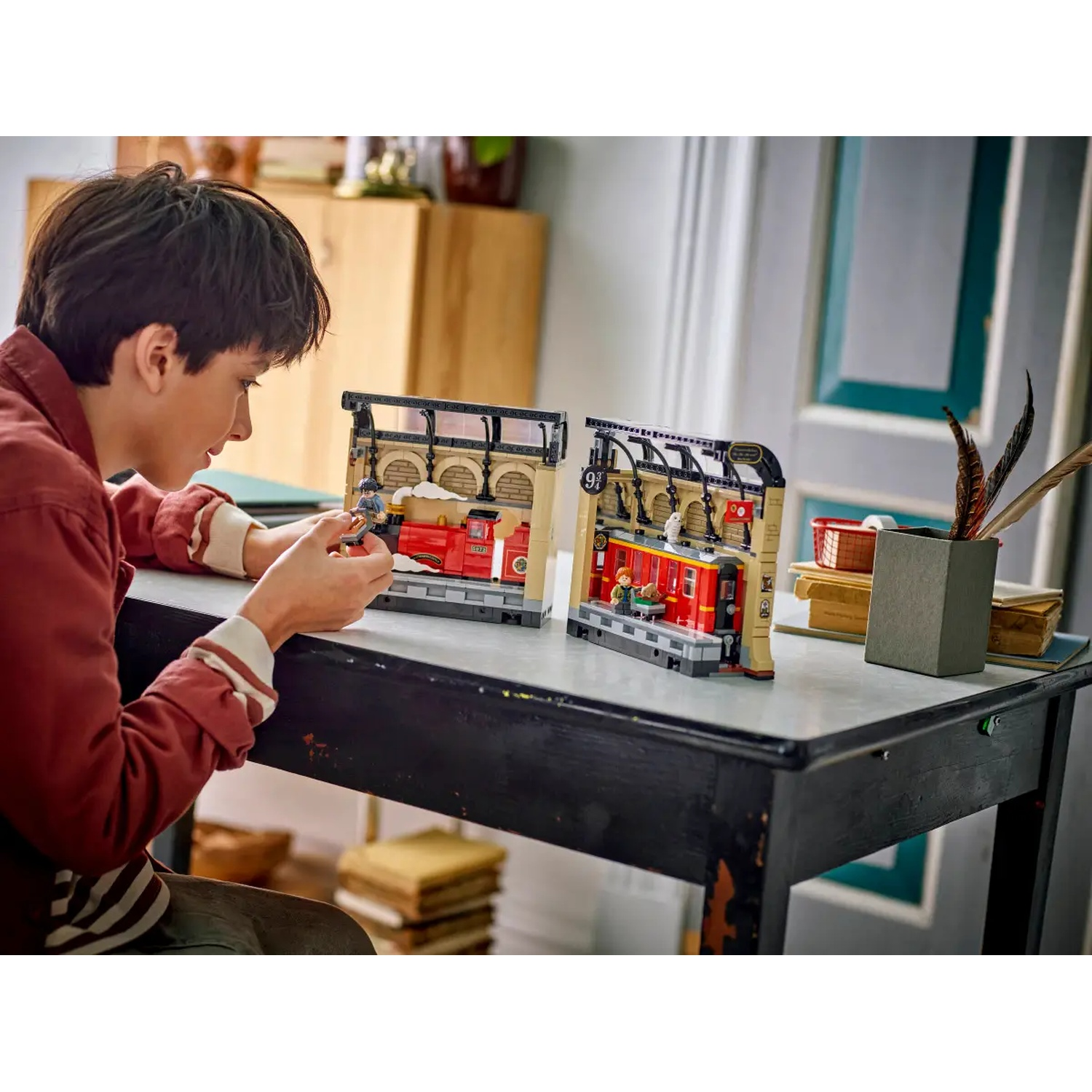 A person is assembling a Lego Harry Potter set. The set depicts a train station with a red train. A desk holds the set pieces.