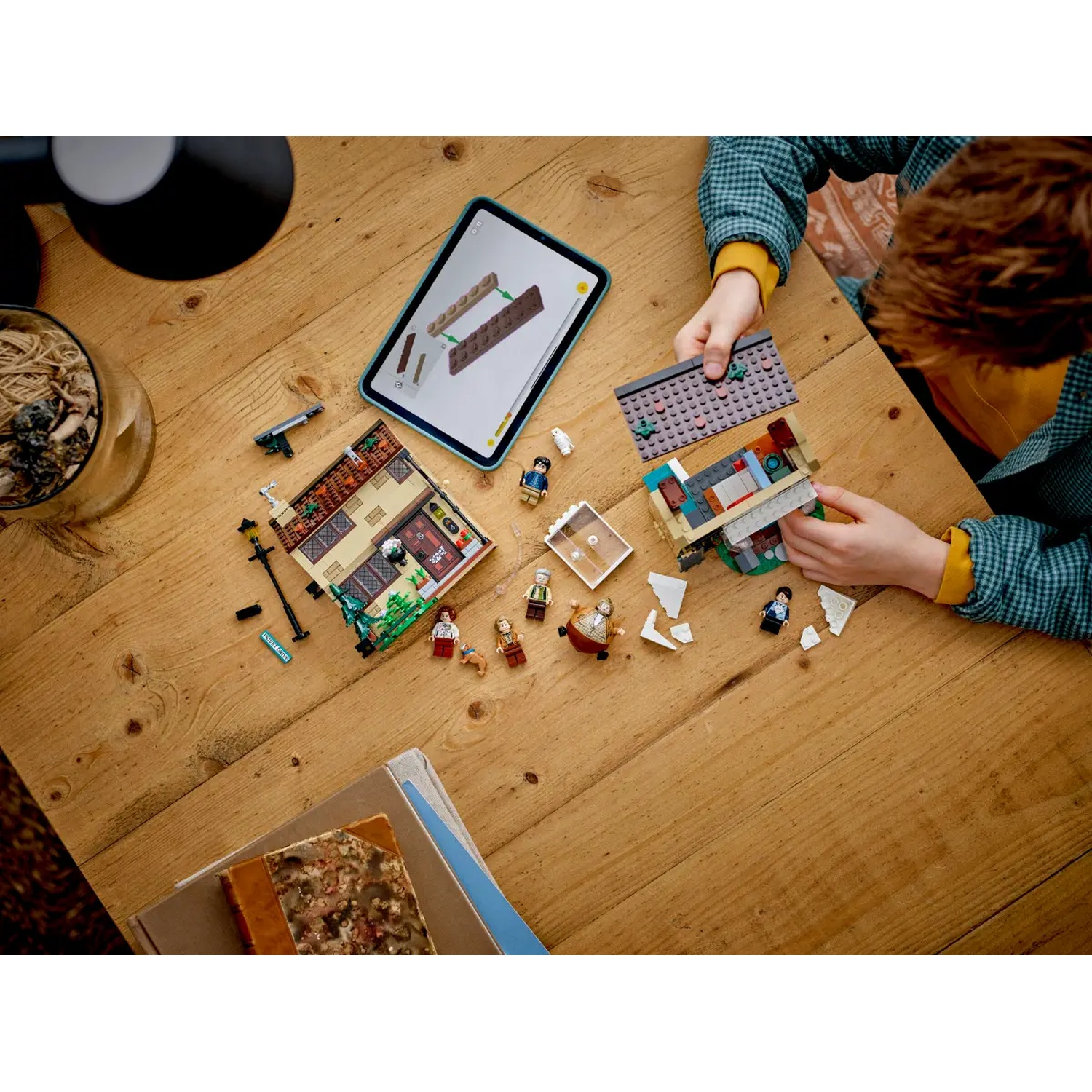 A child is building a Lego house on a wooden table. A tablet shows instructions. Lego figures and pieces are scattered around.